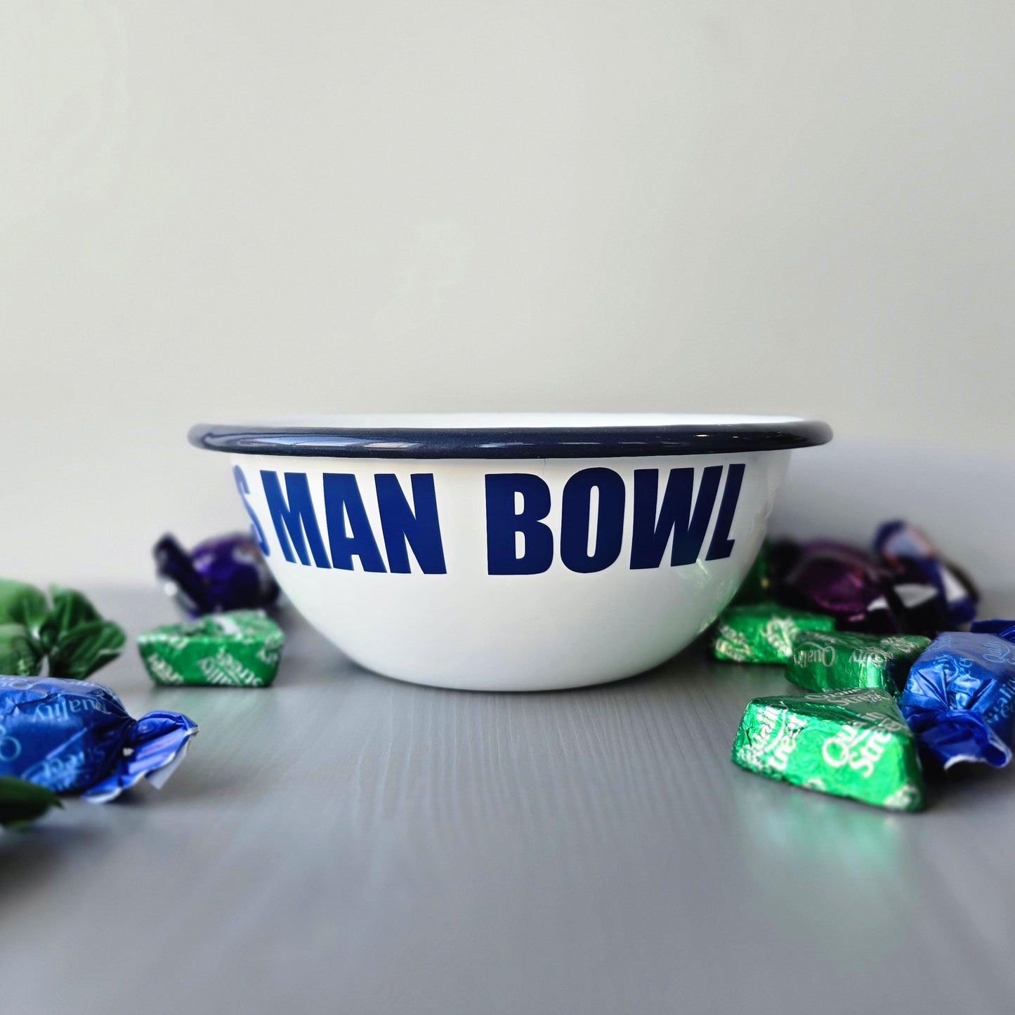 White enamel metal bowl with blue rim and text personalised with a name and 'MAN BOWL' on a grey surface.