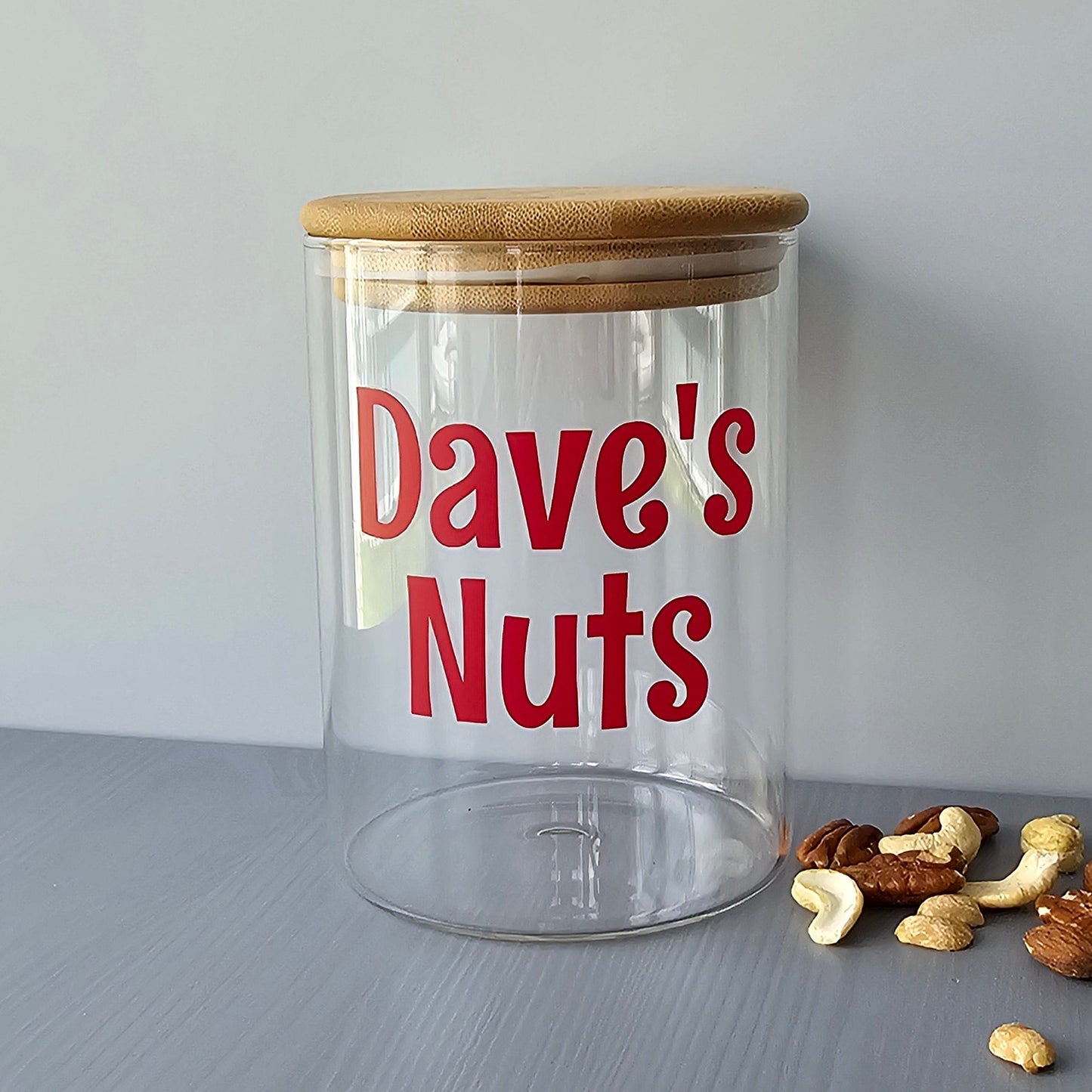 Personalised glass jar with wooden suction lids, labeled 'Dave's Nuts' on a grey surface with nuts around.