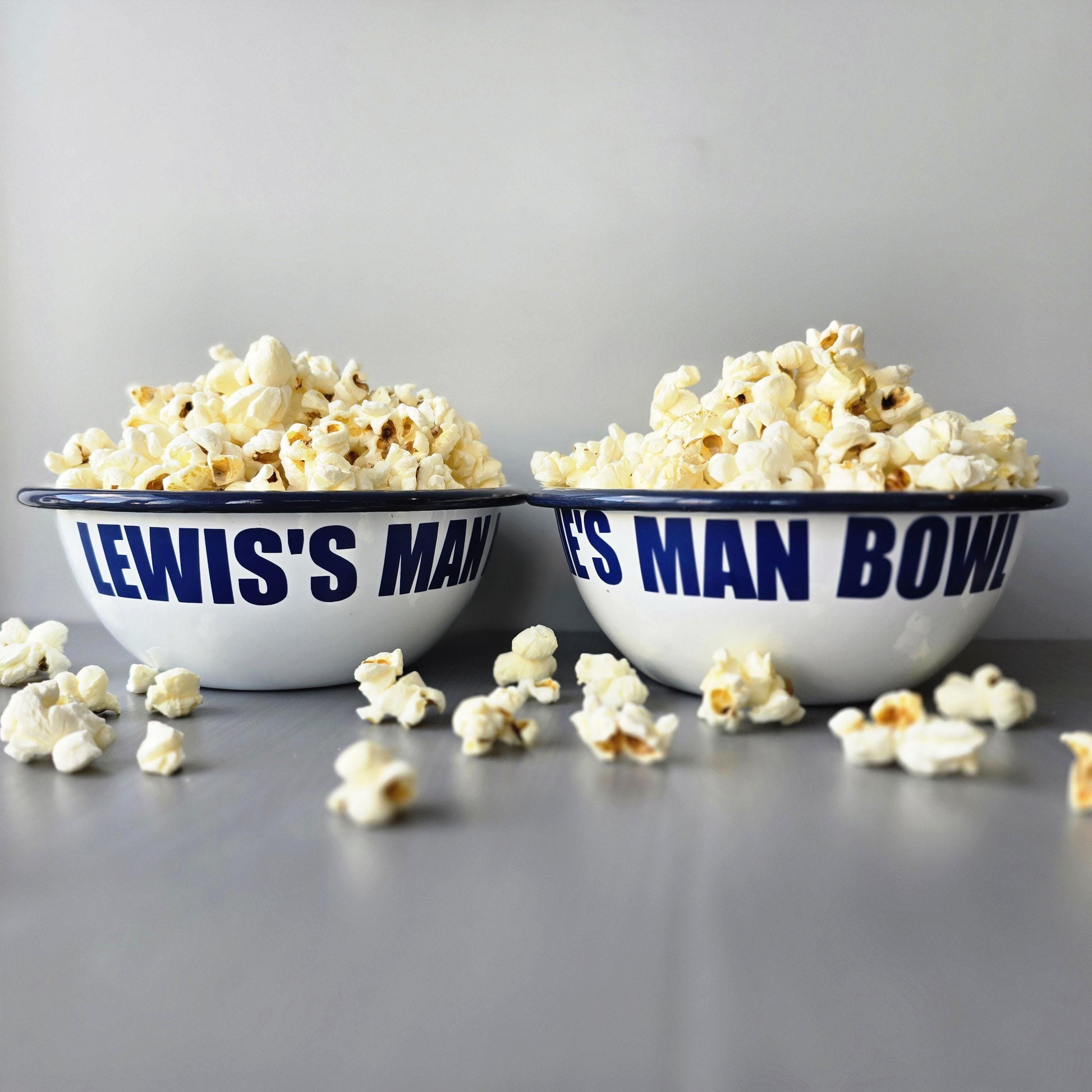 White enamel metal bowl with blue rim and text personalised with a name and 'MAN BOWL' surrounded by popcorn on a grey surface.