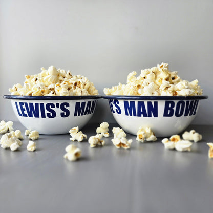 White enamel metal bowl with blue rim and text personalised with a name and 'MAN BOWL' surrounded by popcorn on a grey surface.