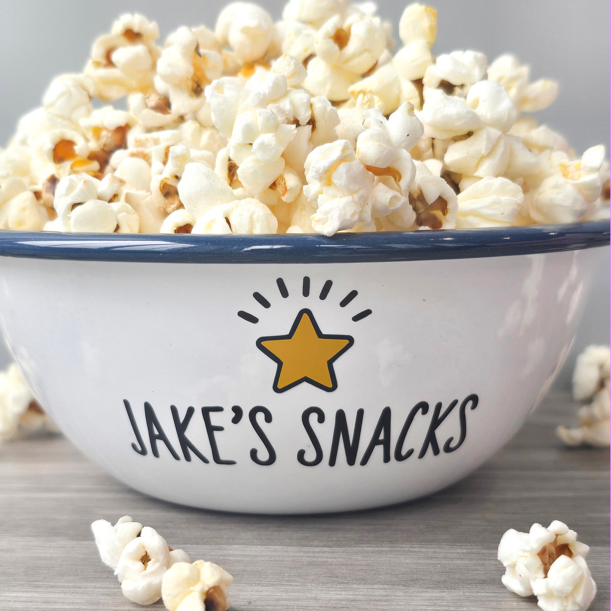 Kids Personalised white metal enamel snack bowl. White enamel metal bowl with navy blue rim. Printed with a yellow star or red heart and a name with snacks written underneath. Perfect for popcorn, sweets, crisps or general snacks.