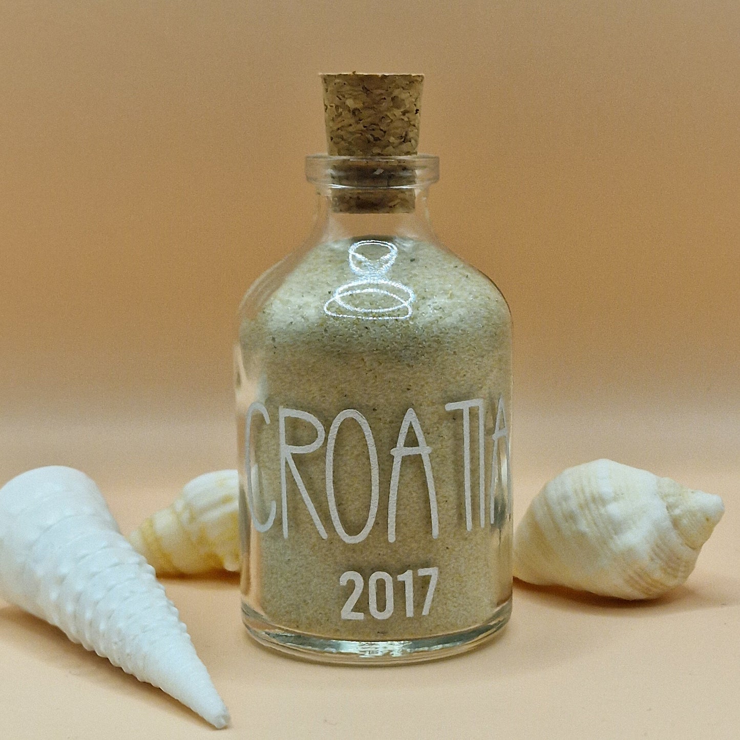 Glass bottle filled with sand labeled 'Croatia 2017' with shells on a beige background