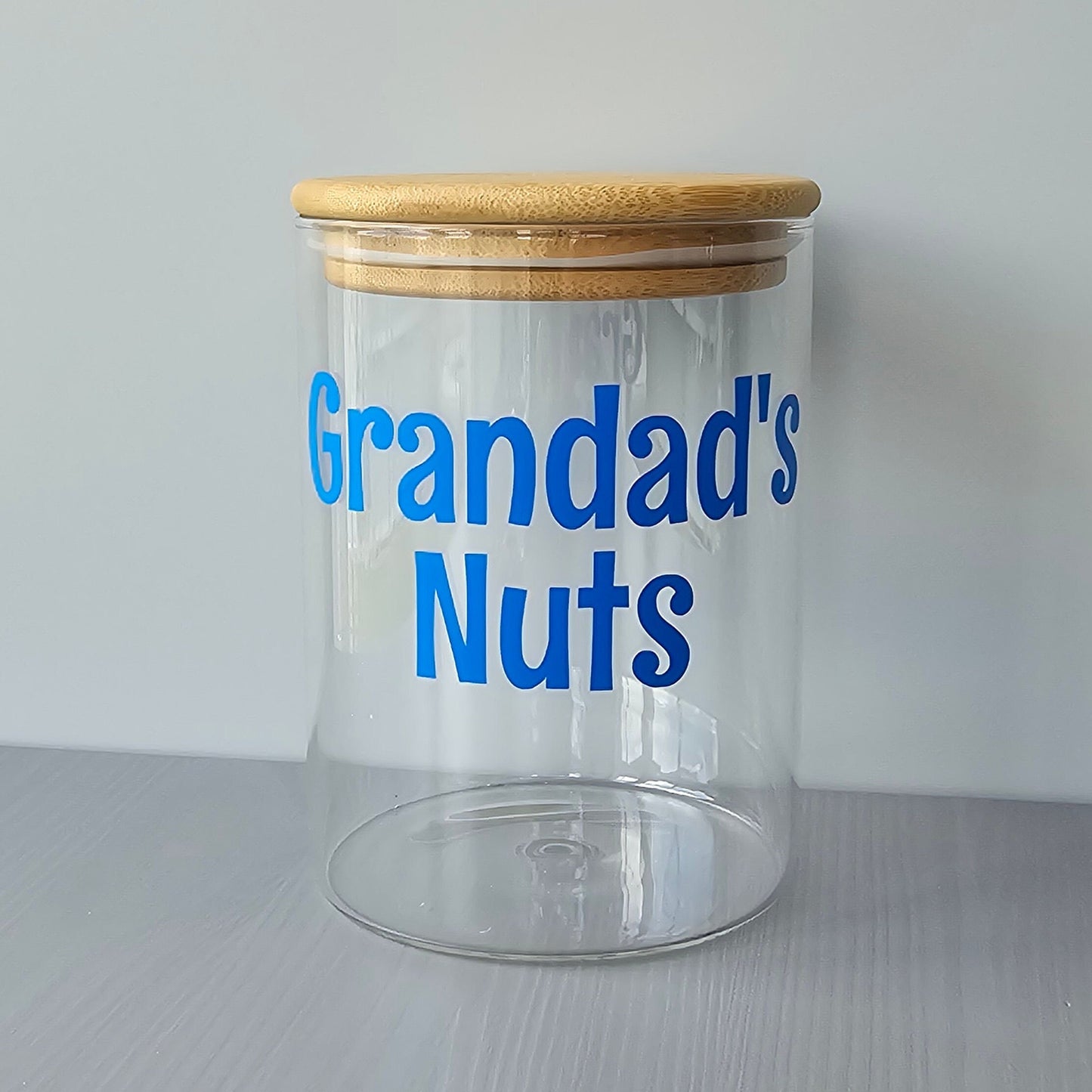 Personalised glass jar with wooden suction lids and blue text 'Grandad's Nuts' on a grey surface.
