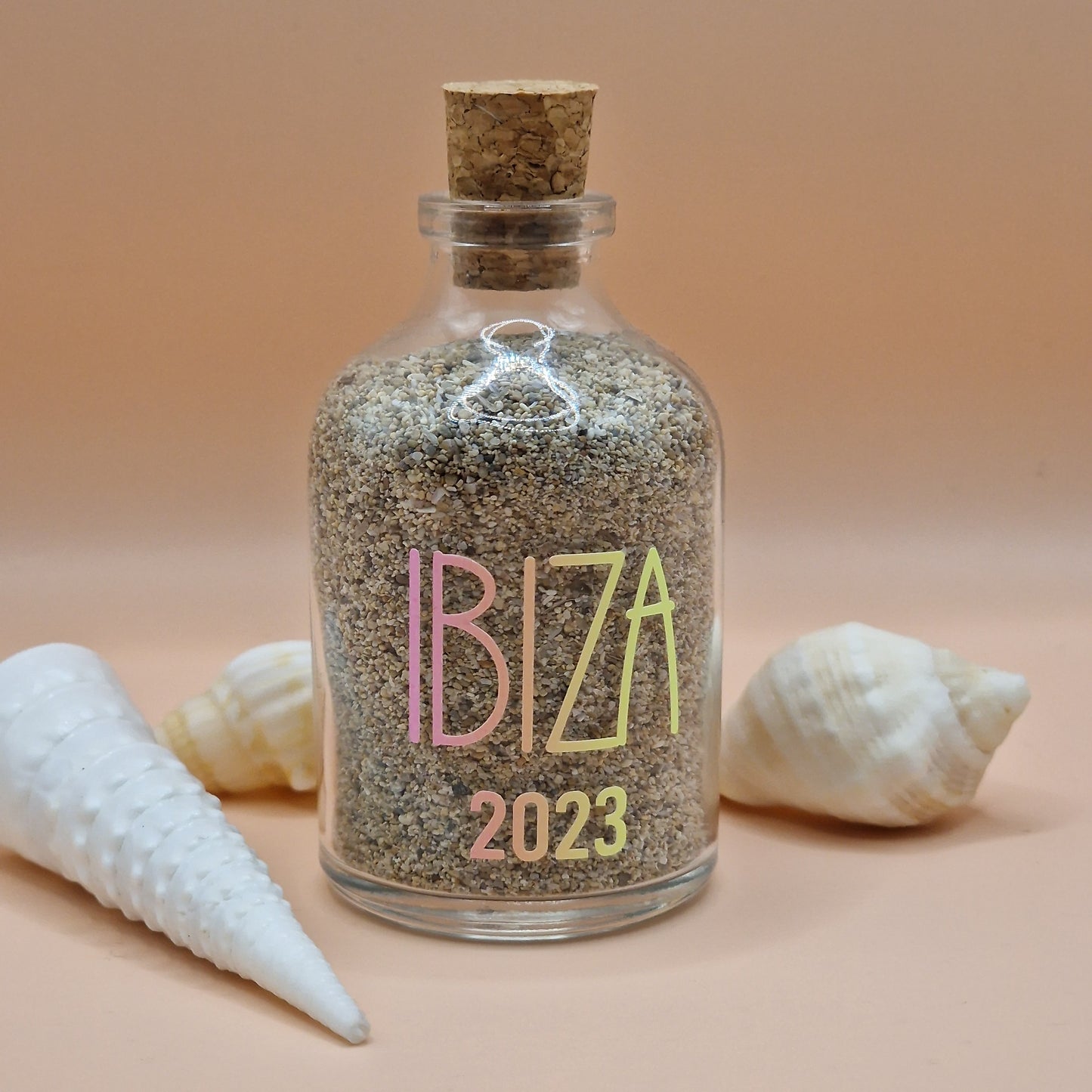 Bottle filled with sand and 'IBIZA 2023' text, surrounded by shells on a peach background