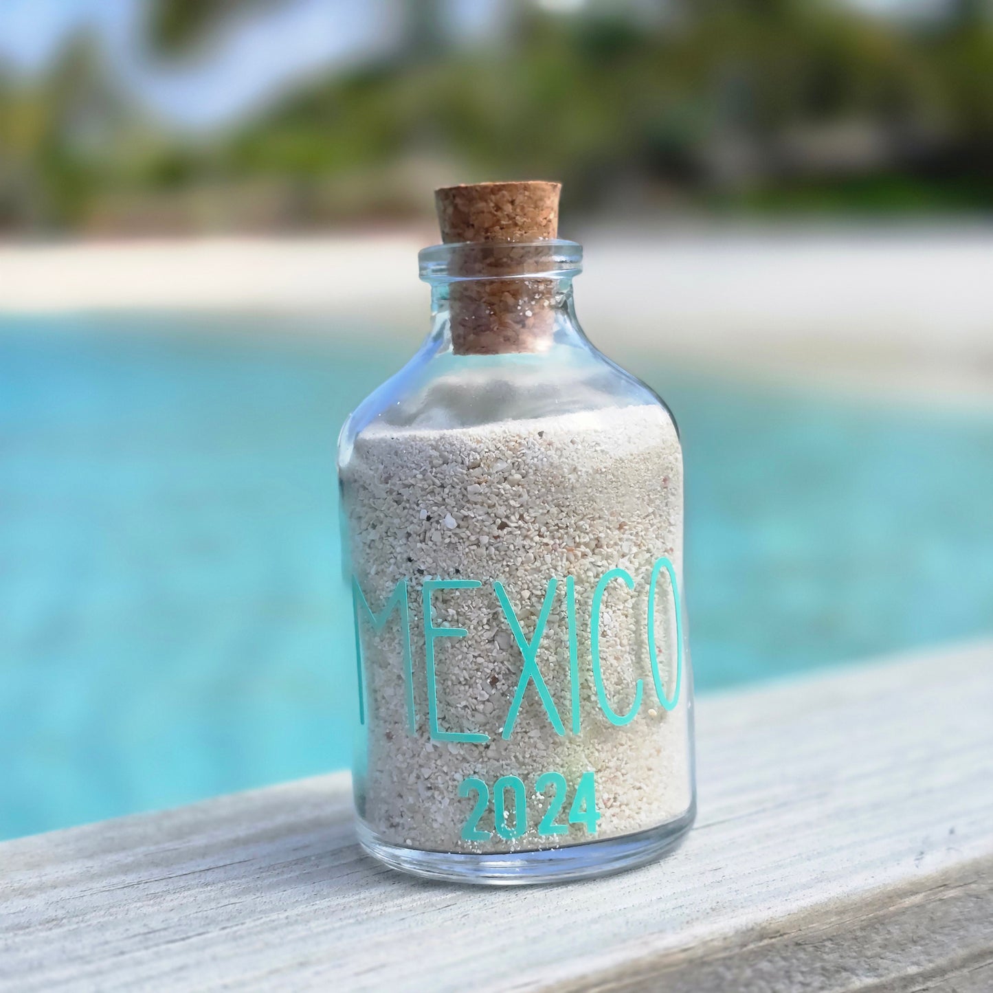 Glass bottle with sand and 'MEXICO 2024' text against a blurred natural background