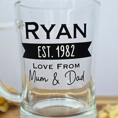 Glass beer tankard personalised with a name, year and signed from the gift giver in black vinyl on clear glass. Sturdy, heavy, clear glass stein with mixed font design. 