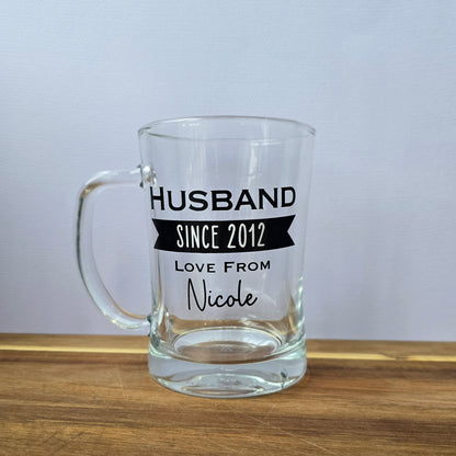 Glass beer tankard personalised with a name, year and signed from the gift giver in black vinyl on clear glass. Sturdy, heavy, clear glass stein with mixed font design. 