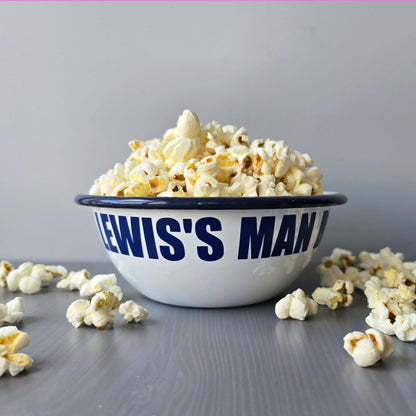 White enamel metal bowl with blue rim and text personalised with a name and 'MAN BOWL' surrounded by popcorn on a grey surface.