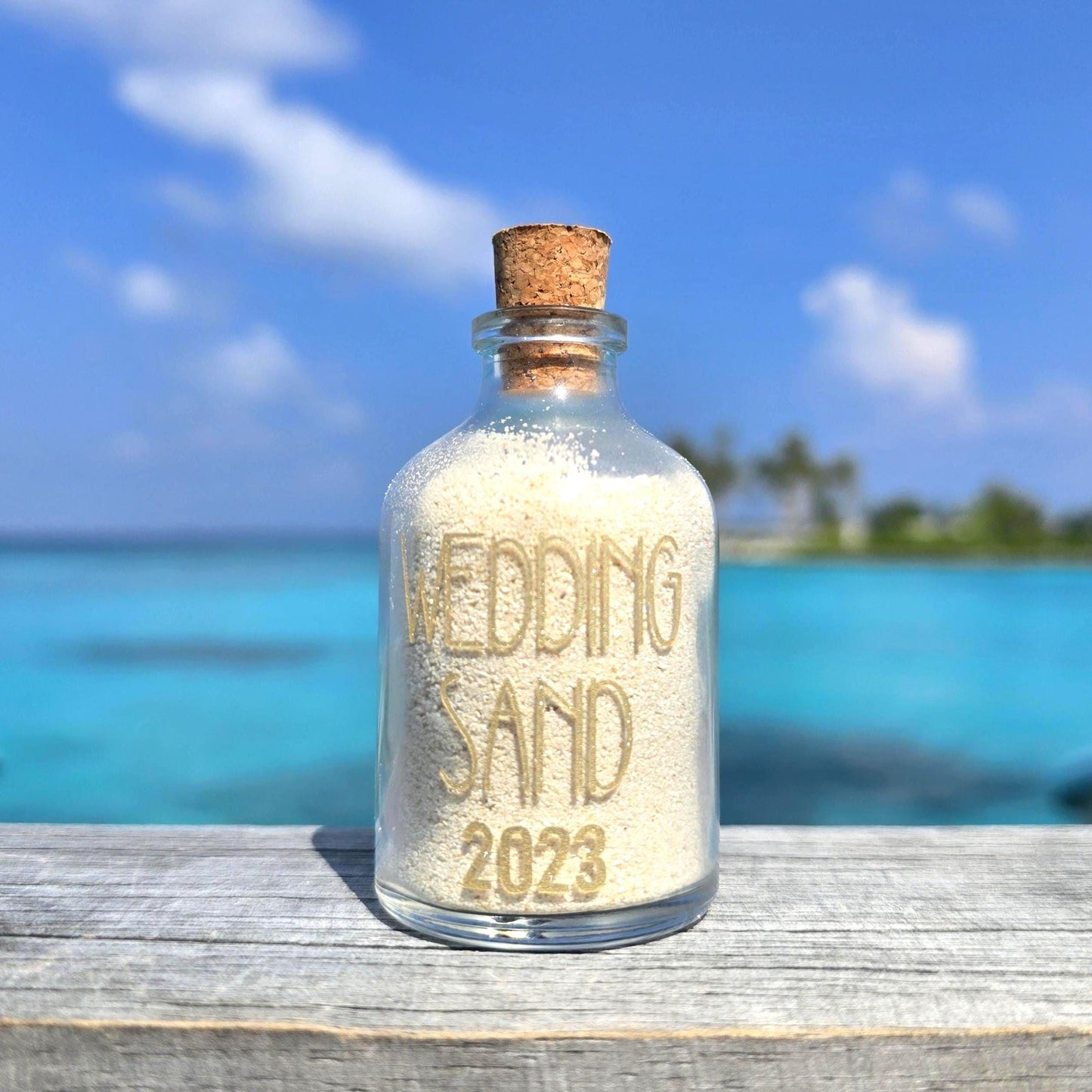 Glass bottle labeled 'Wedding Sand 2023' with a cork lid on a wooden surface with a beach background.