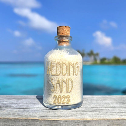 Glass bottle labeled 'Wedding Sand 2023' with a cork lid on a wooden surface with a beach background.
