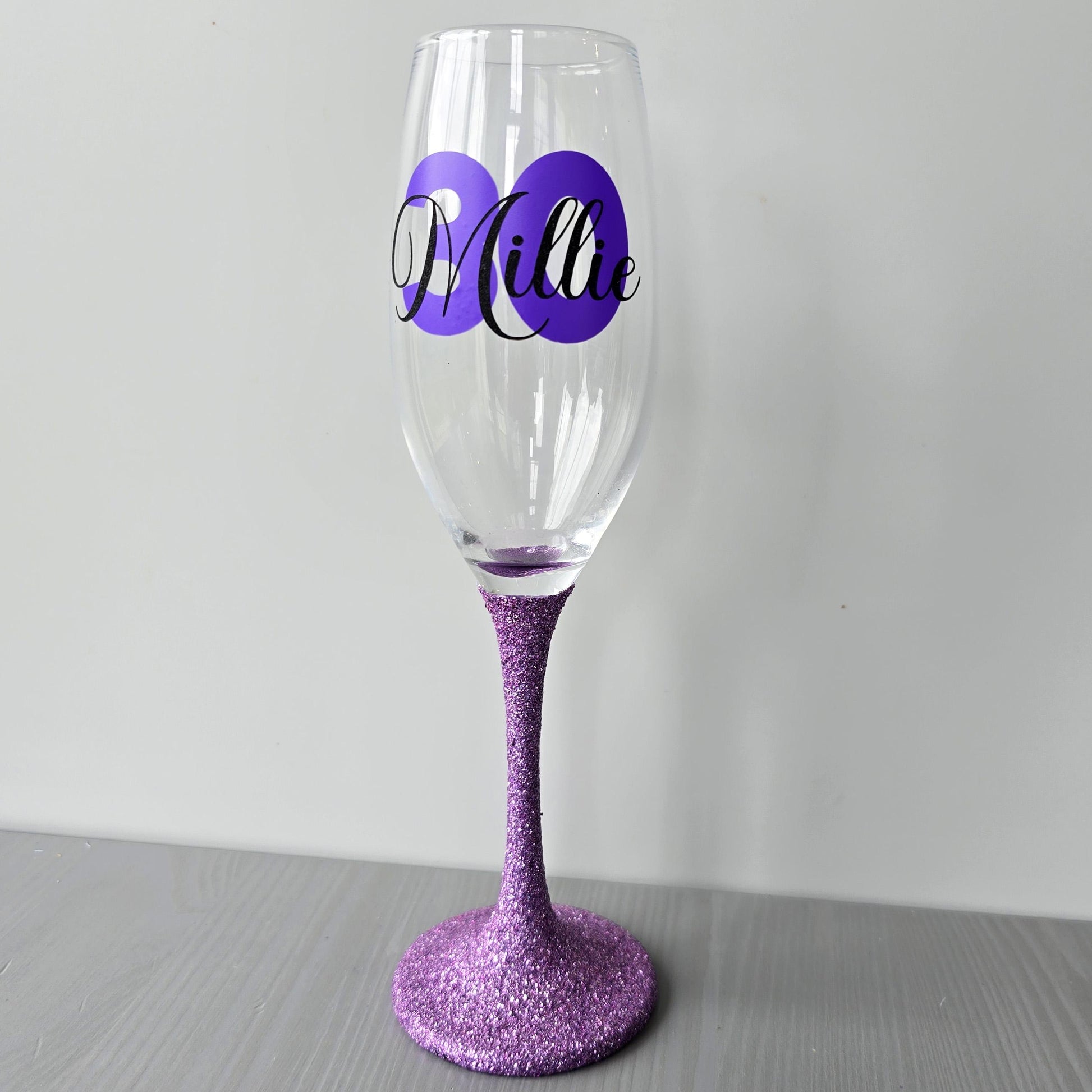 Personalised champagne flute or prosecco glass with glitter stem and with a birthday age number in block, glitter or metallic vinyl and a name in black decorative font through the number