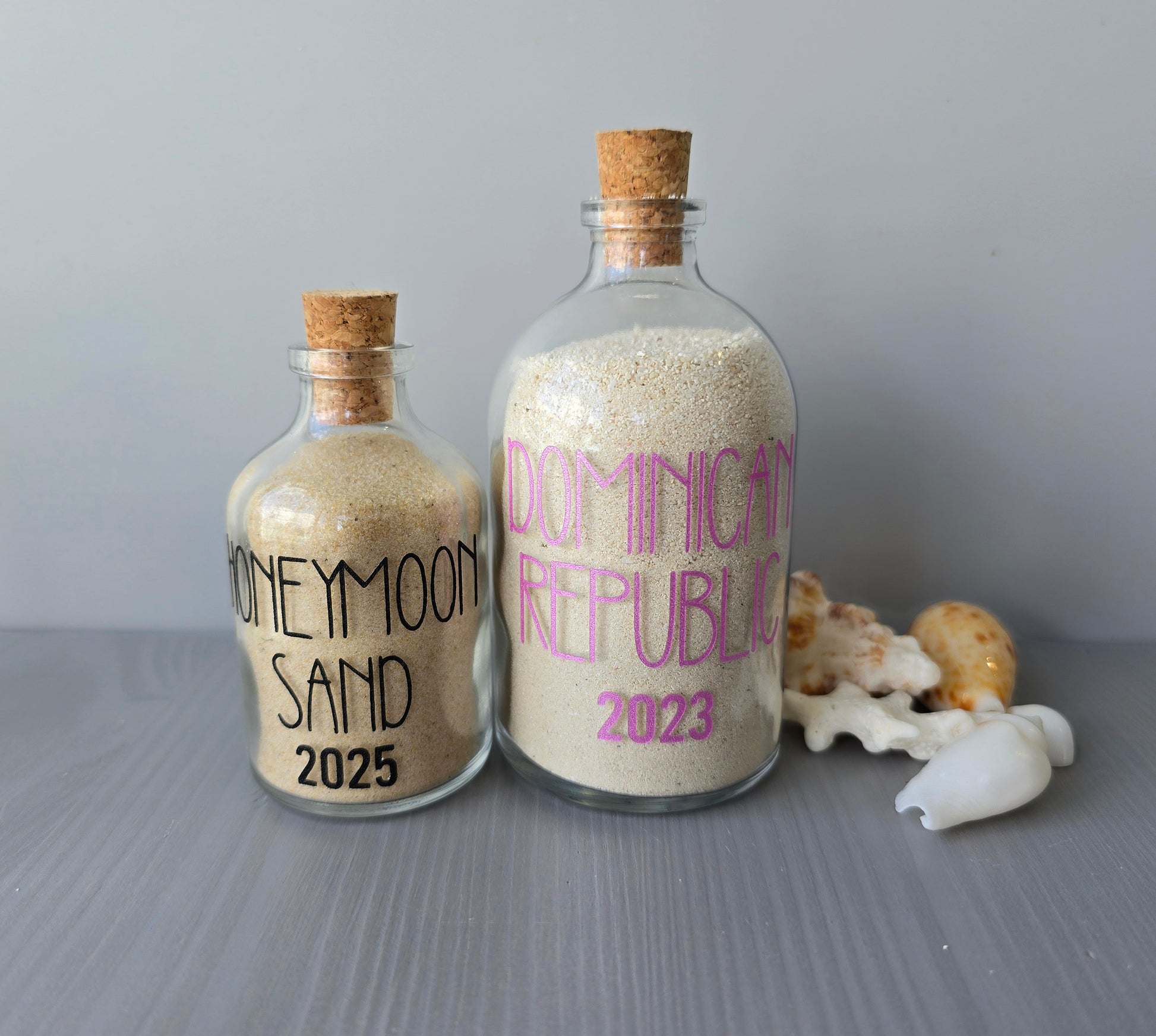 Two bottles filled with sand labeled 'Honeymoon Sand 2025' and 'Dominican Republic 2023' on a gray surface.