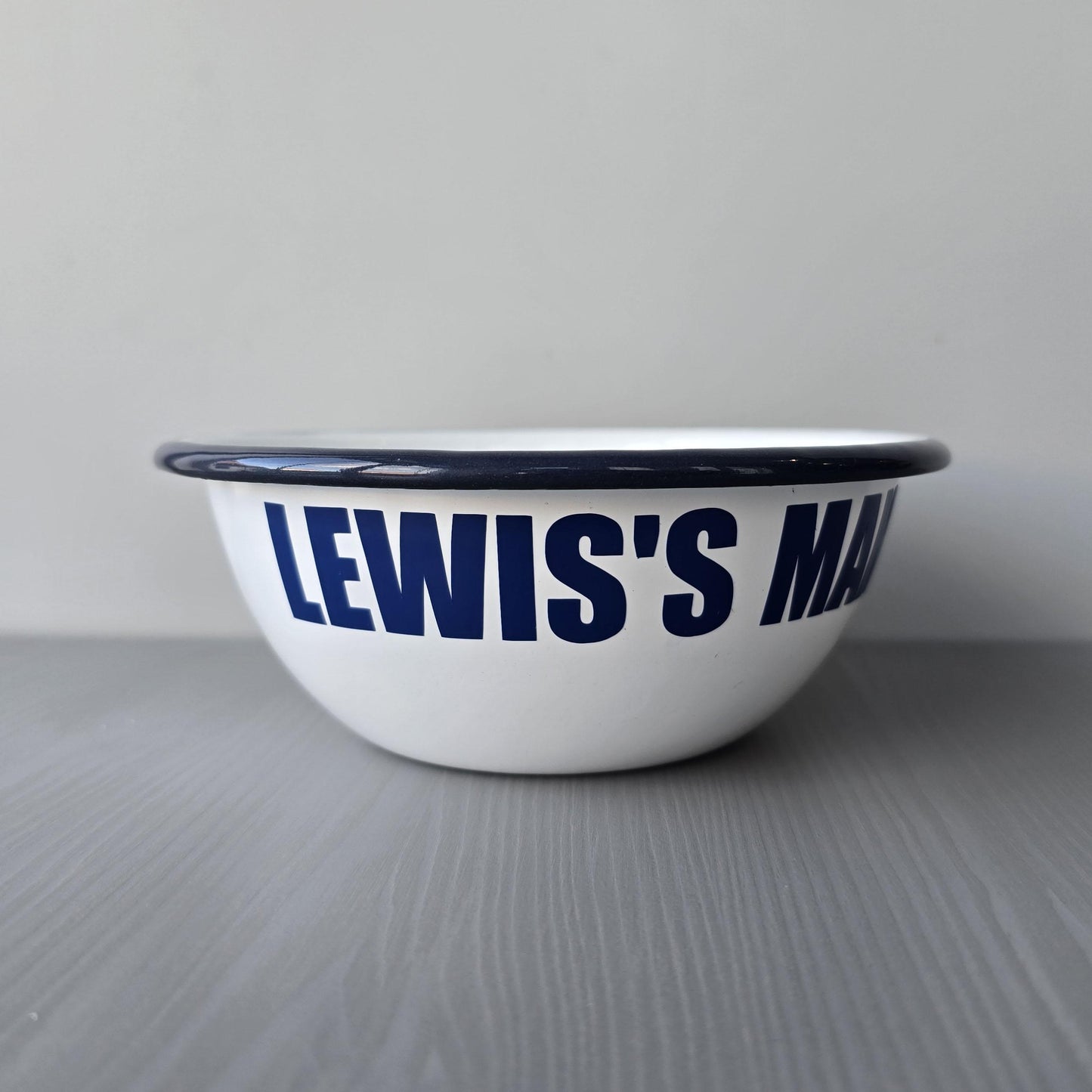 White enamel metal bowl with blue rim and text personalised with a name and 'MAN BOWL' on a grey surface.