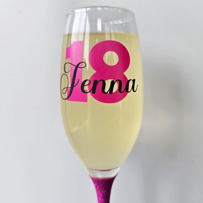 Personalised champagne flute or prosecco glass with glitter stem and with a birthday age number in block, glitter or metallic vinyl and a name in black decorative font through the number