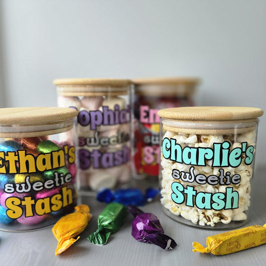 Personalised glass jars with wodden suction lid  labeled 'Ethan's sweetie Stash' and 'Charlie's sweetie Stash' with colourful candy wrappers on a gray background.