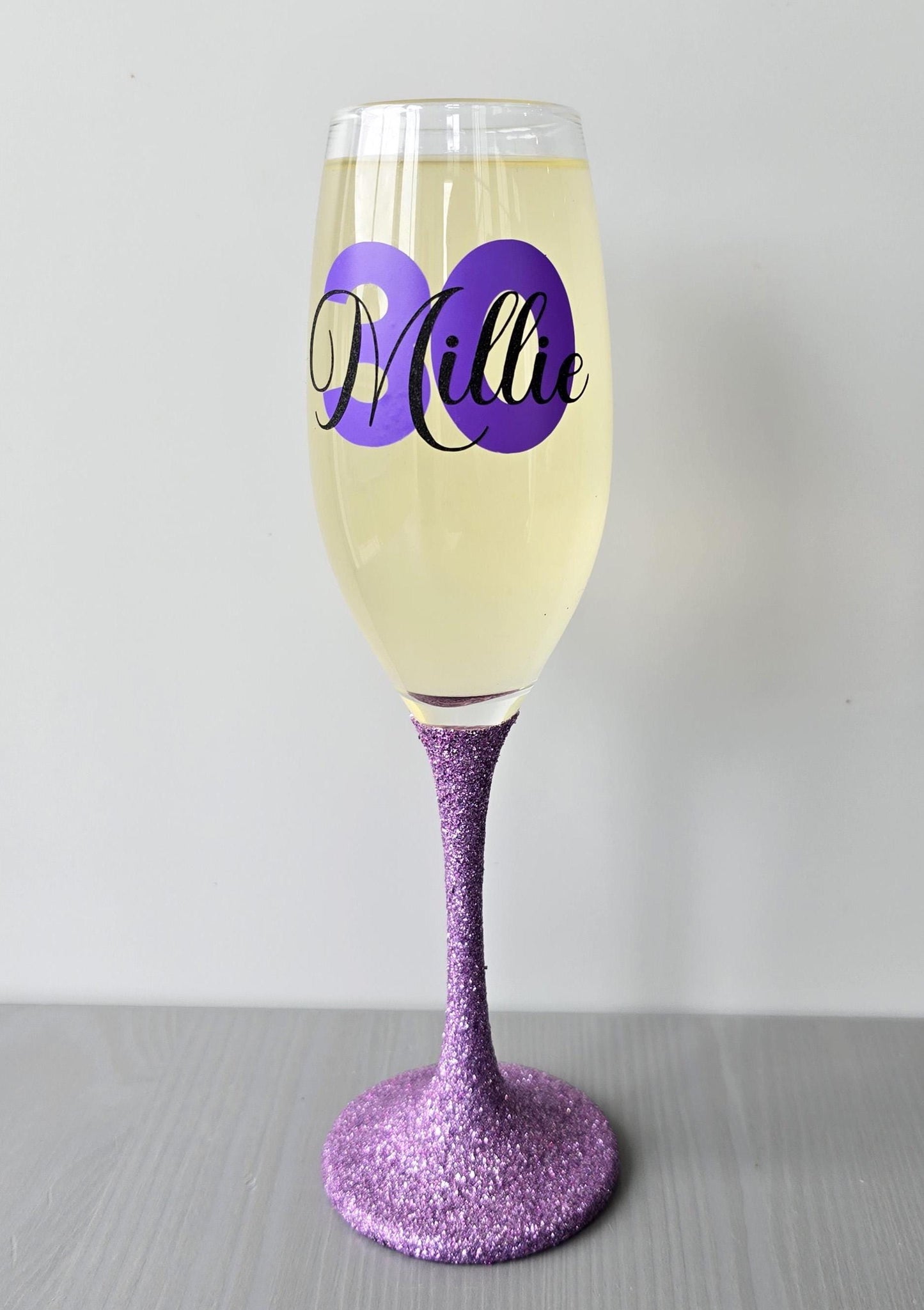 Personalised champagne flute or prosecco glass with glitter stem and with a birthday age number in block, glitter or metallic vinyl and a name in black decorative font through the number