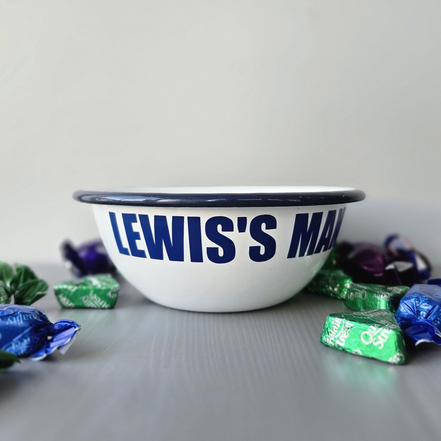 White enamel metal bowl with blue rim and text personalised with a name and 'MAN BOWL' on a grey surface.