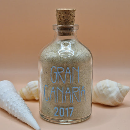 Glass bottle filled with sand labeled 'Gran Canaria 2017' with shells on a beige background