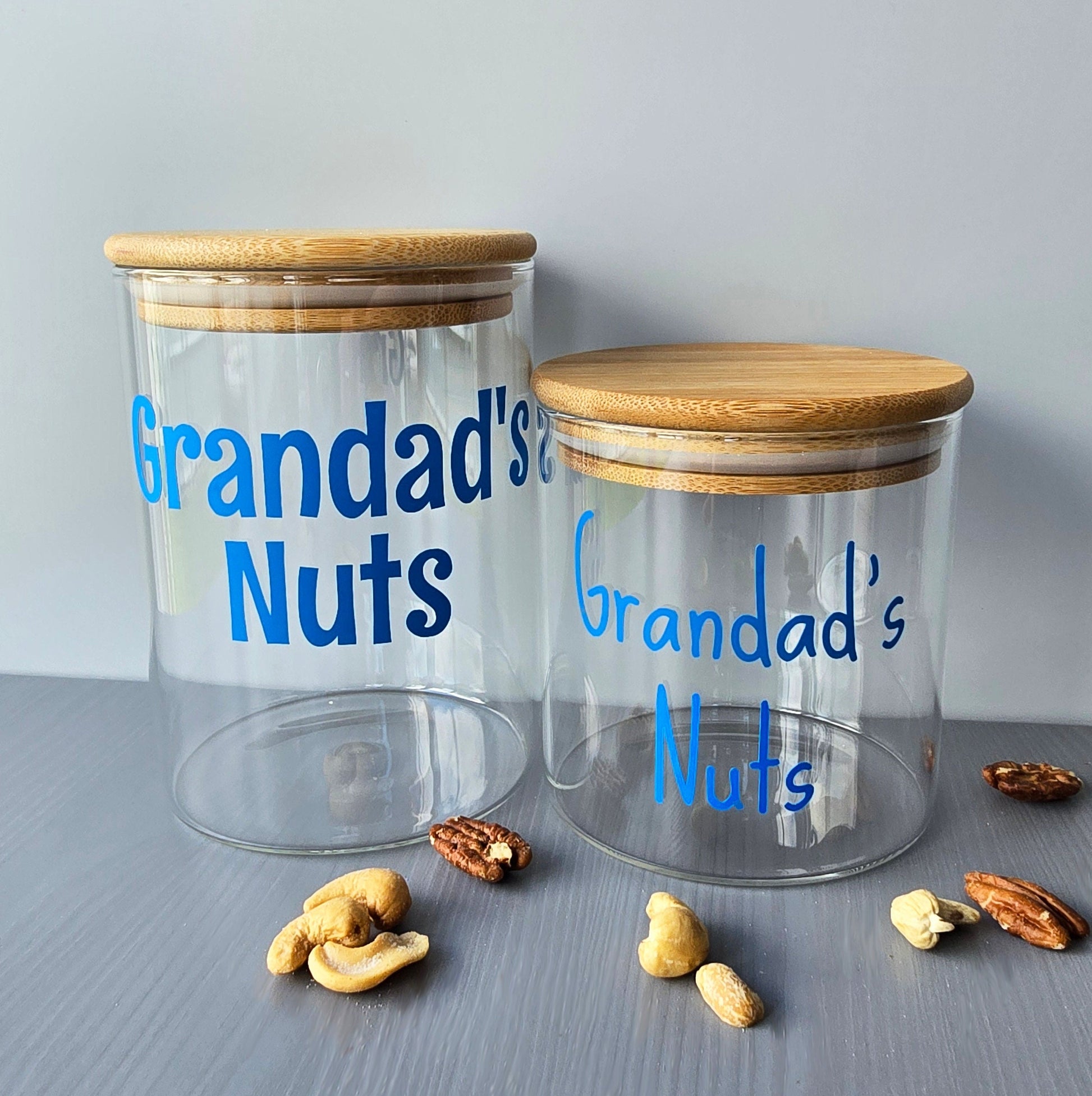 Two glass jars with wooden suction lids labeled 'Grandad's Nuts' on a grey surface with nuts around.