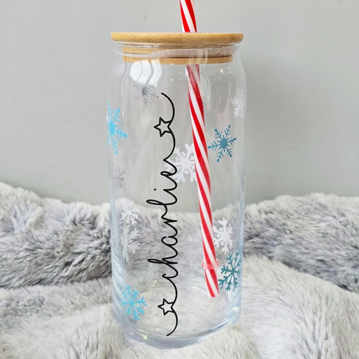 Personalised glass can drink holder with a bamboo suction lid and red and white reusable straw. Features silver snowflakes and blue snowflakes that colour change to purple when cold liquid is added to the glass. Personalised name along the side of the glass, available in glittery silver or black and features either a heart or stars at either end of the name.