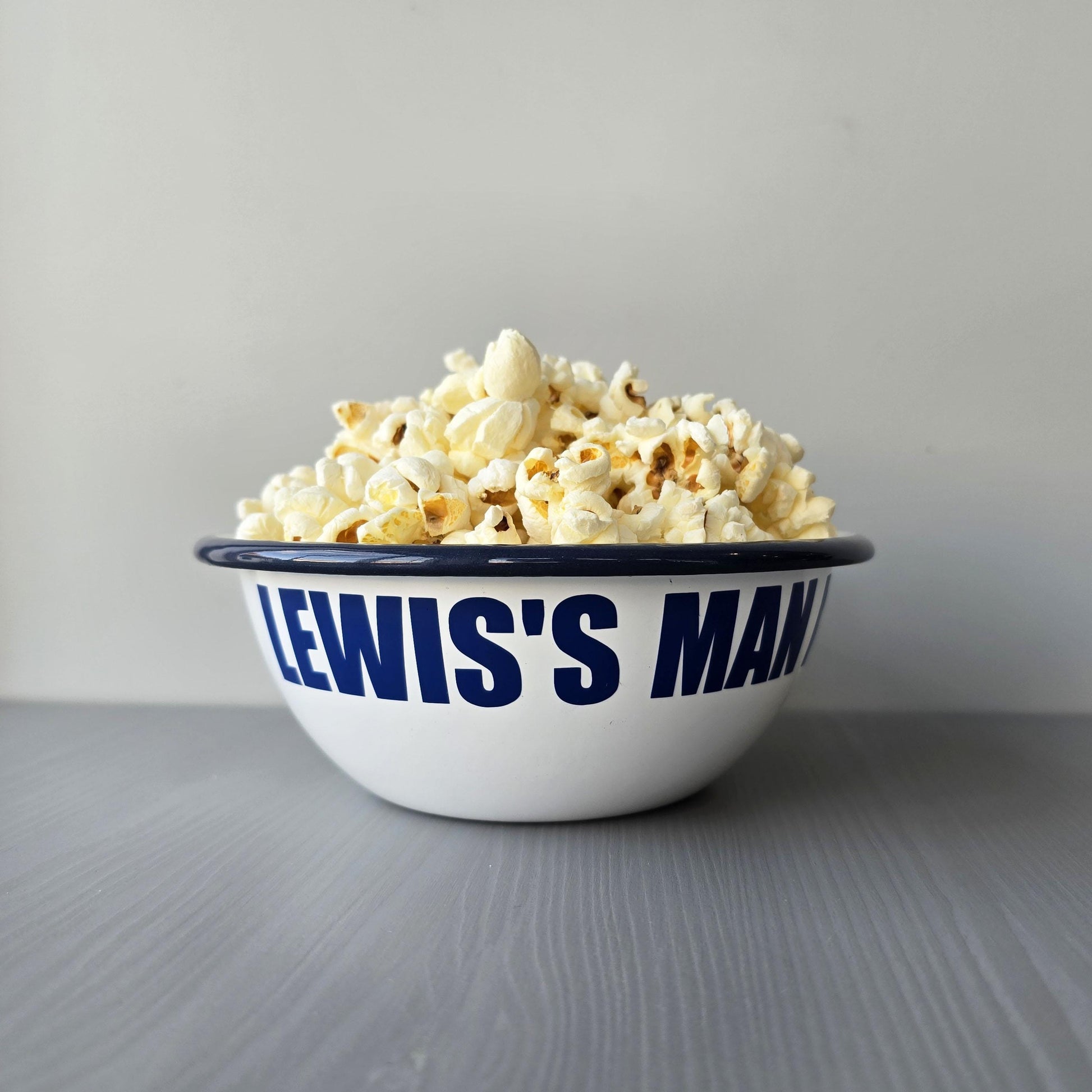 White enamel metal bowl with blue rim and text personalised with a name and 'MAN BOWL' surrounded by popcorn on a grey surface.
