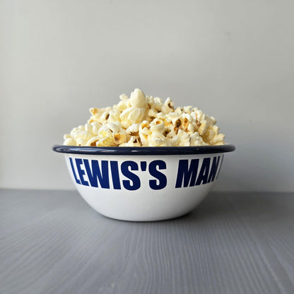 White enamel metal bowl with blue rim and text personalised with a name and 'MAN BOWL' surrounded by popcorn on a grey surface.