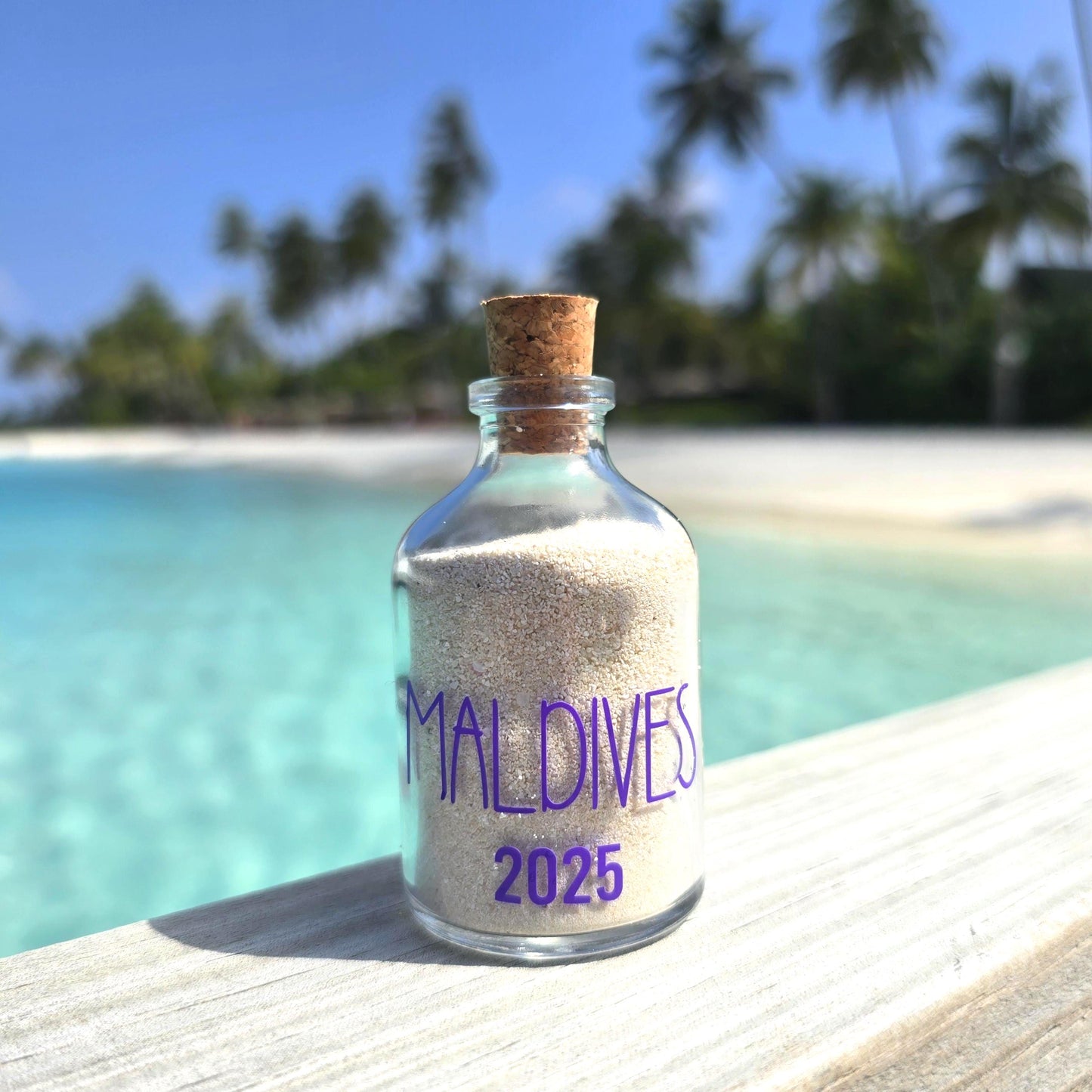 Glass bottle with cork lid containing sand labeled 'Maldives 2025' on a wooden surface with a tropical beach background.