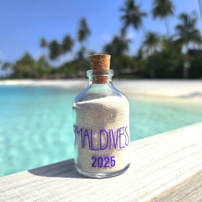 Glass bottle with cork lid containing sand labeled 'Maldives 2025' on a wooden surface with a tropical beach background.
