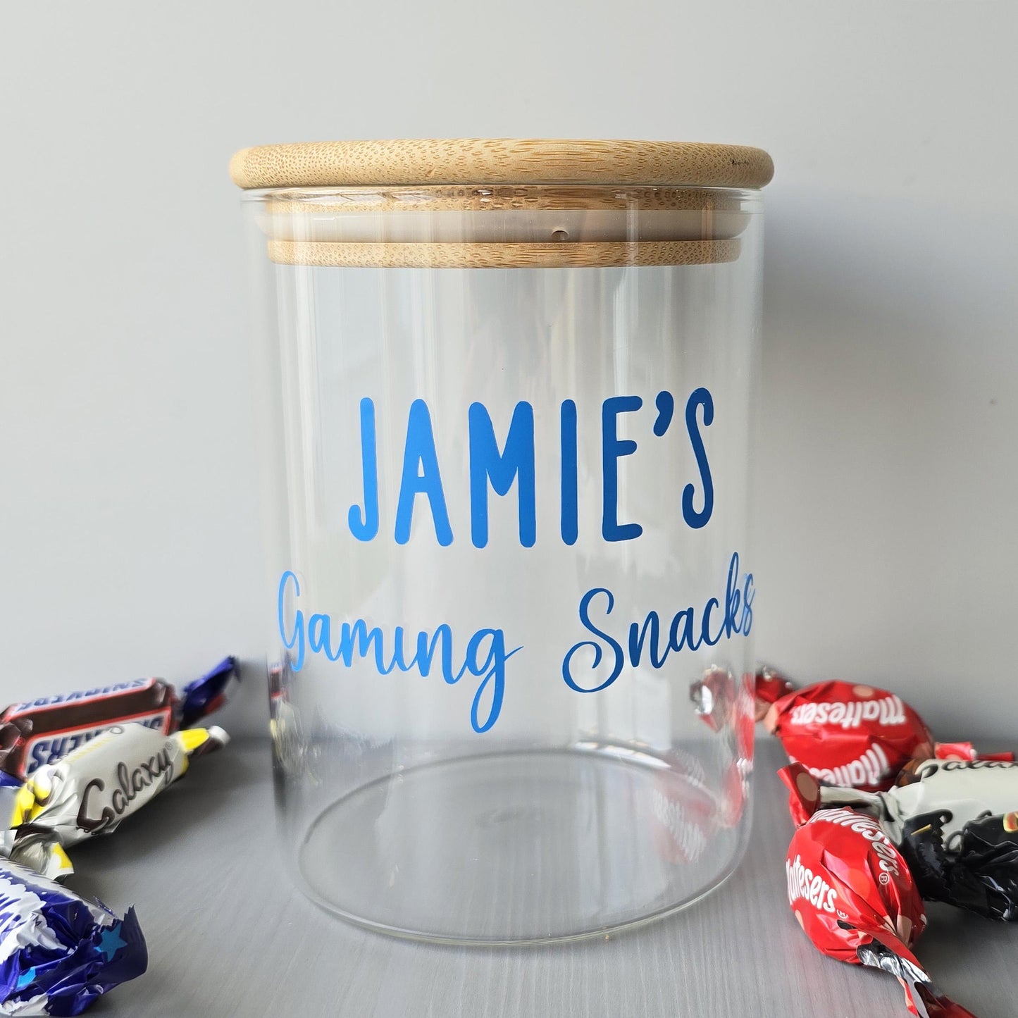 Clear jar with wooden lid labeled 'Jamie's Gaming Snacks' on a gray background with candy bars.