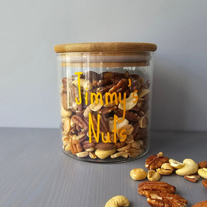 Personalised glass jar with wooden suction lids, labeled 'Jimmy's Nuts' filled with mixed nuts on a grey surface.