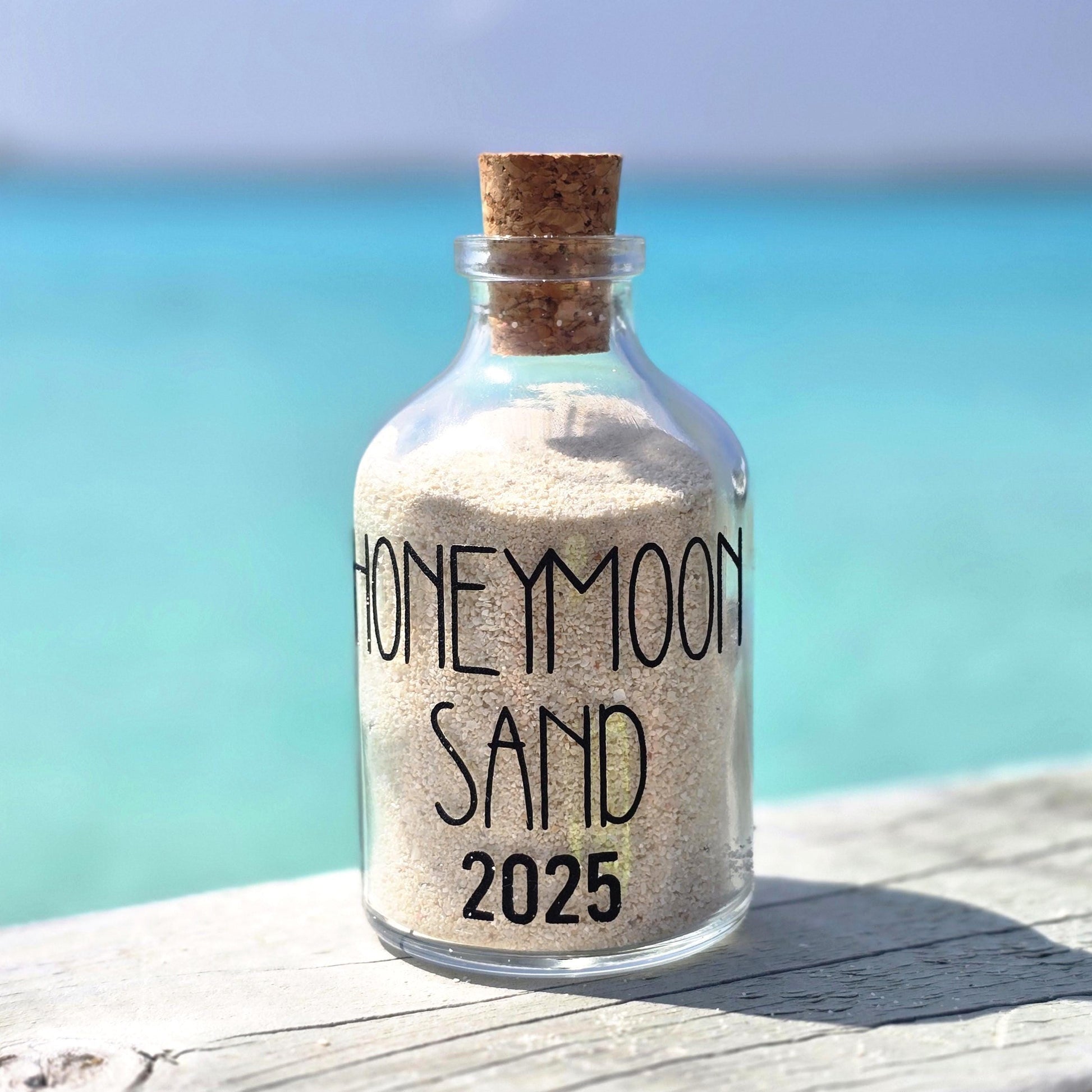 Glass bottle labeled 'Honeymoon Sand 2025' filled with sand, set against a beach background.