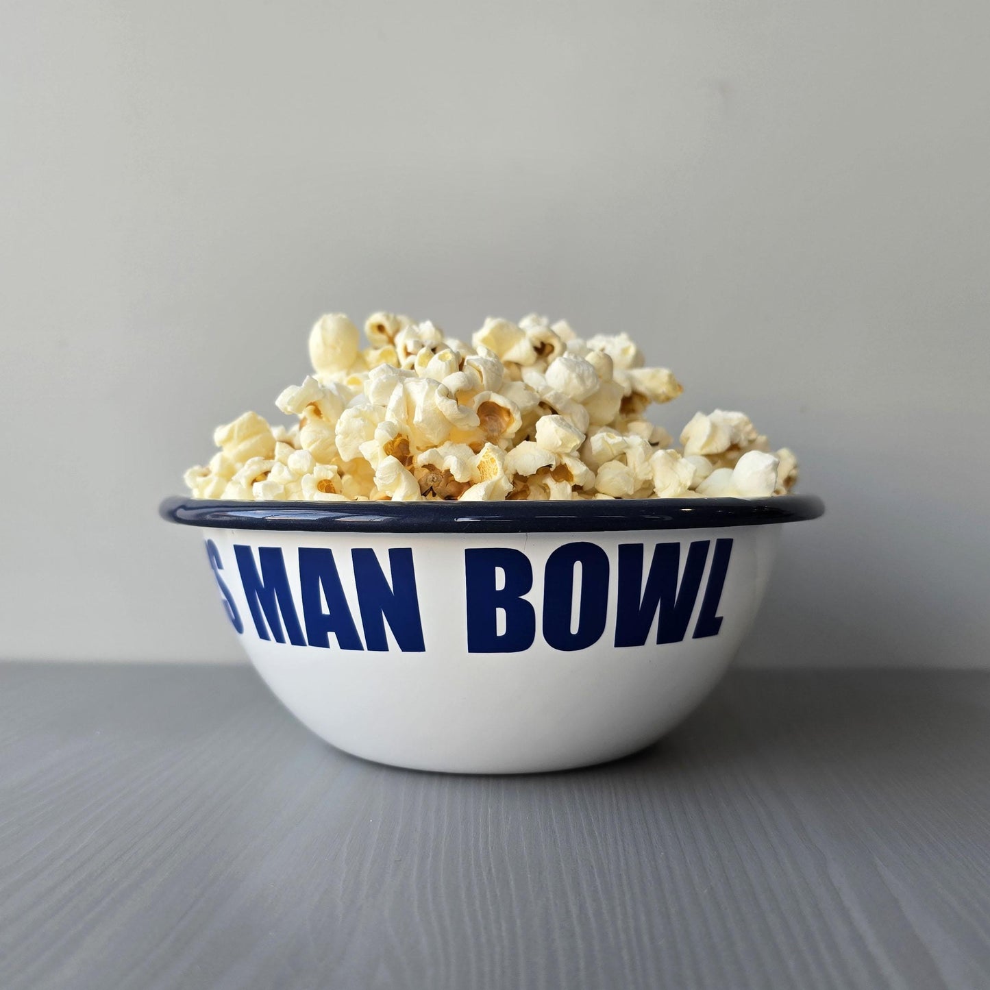 White enamel metal bowl with blue rim and text personalised with a name and 'MAN BOWL' surrounded by popcorn on a grey surface.
