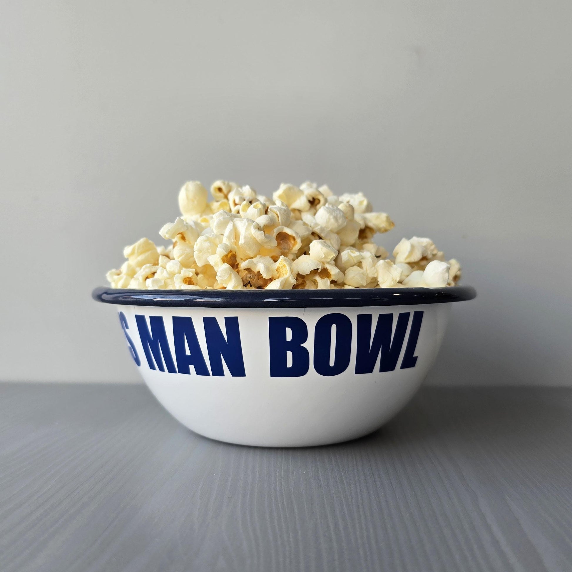 White enamel metal bowl with blue rim and text personalised with a name and 'MAN BOWL' surrounded by popcorn on a grey surface.