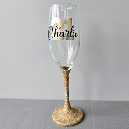 Personalised champagne flute or prosecco glass with glitter stem and with a birthday age number in block, glitter or metallic vinyl and a name in black decorative font through the number