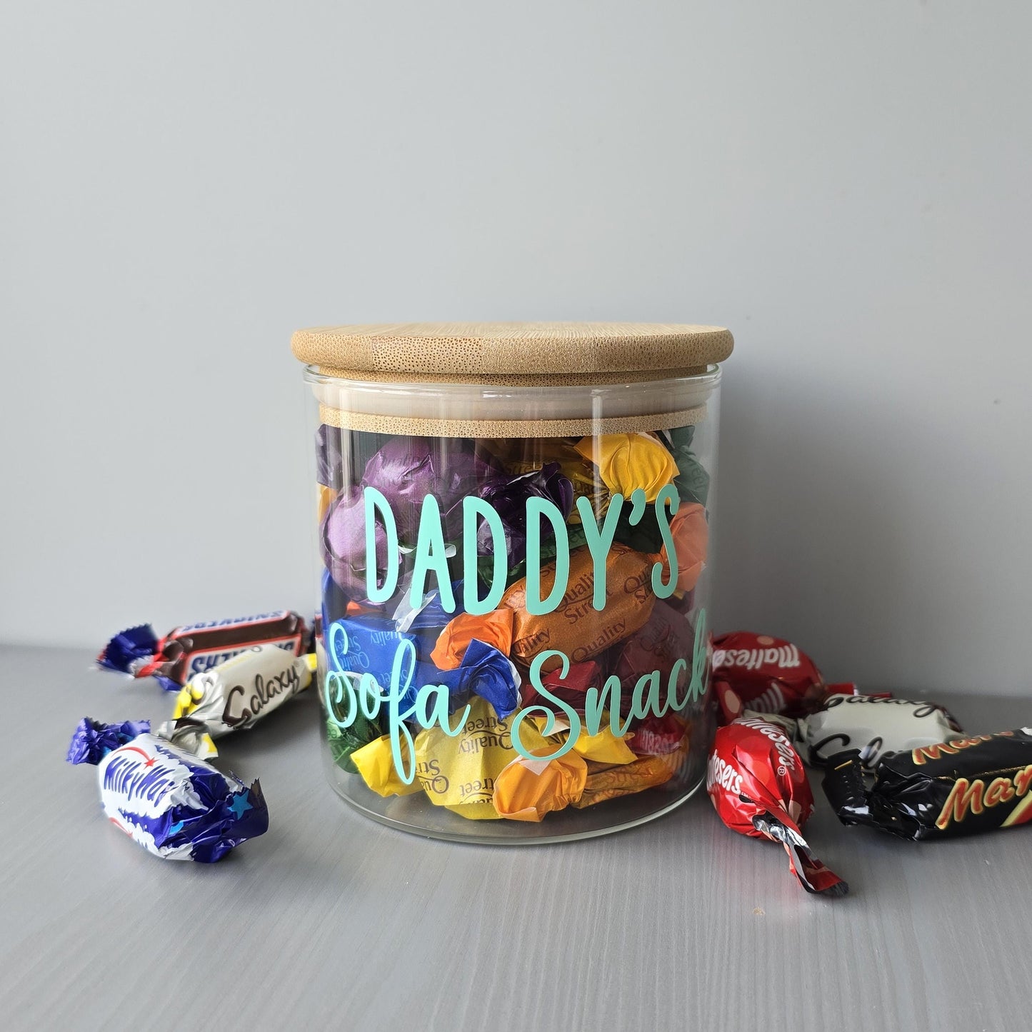 Clear jar labeled 'Daddy's Sofa Snack' filled with assorted candies on a gray surface.