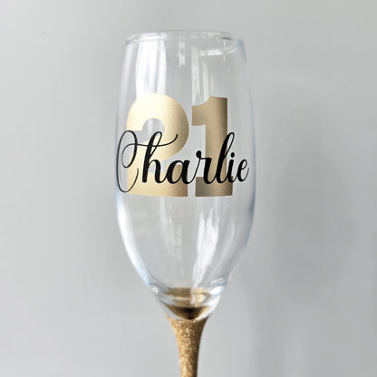 Personalised champagne flute or prosecco glass with glitter stem and with a birthday age number in block, glitter or metallic vinyl and a name in black decorative font through the number