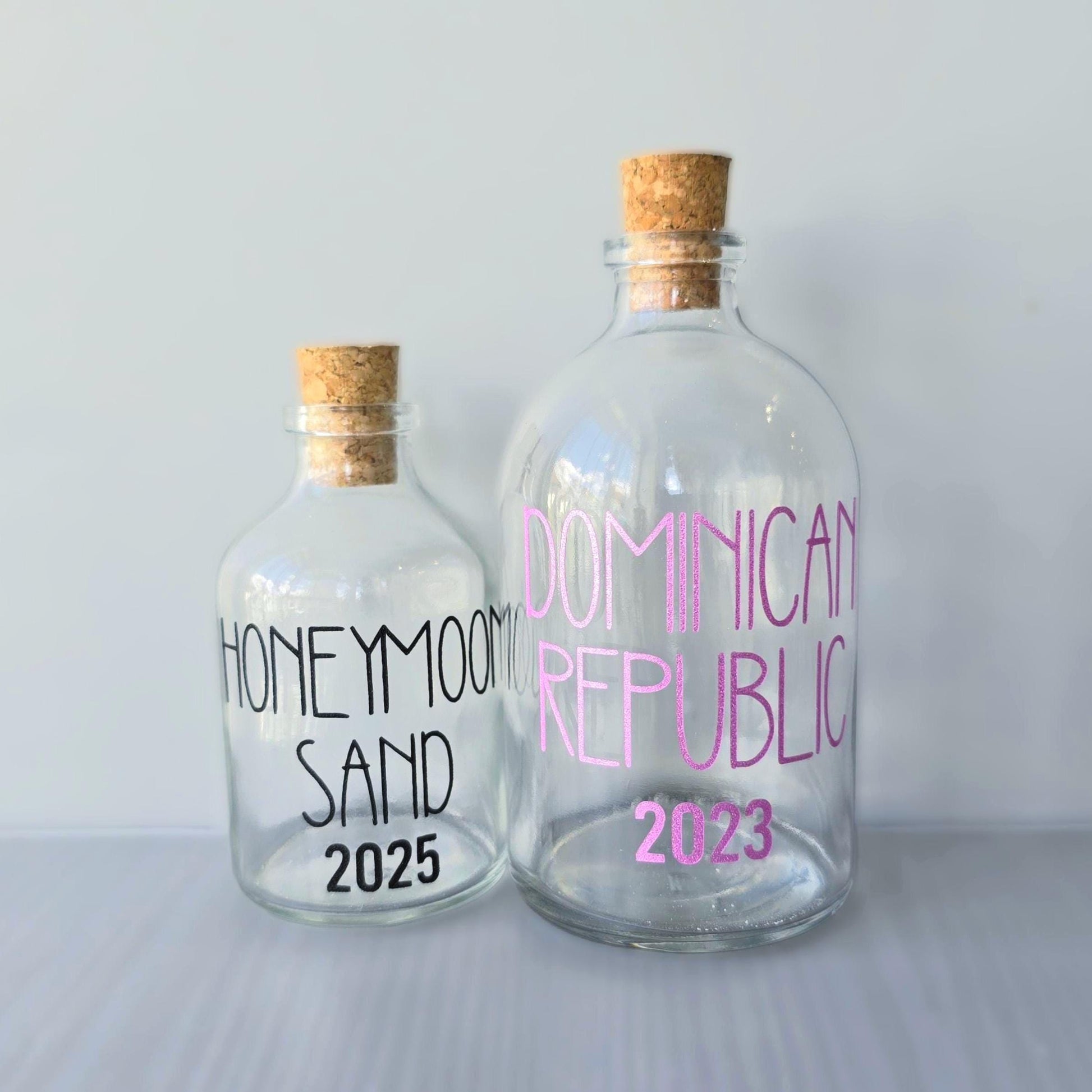 Two glass bottles with cork lids, one labeled 'Honeymoon Sand 2025' and the other 'Dominican Republic 2023', on a light gray background.