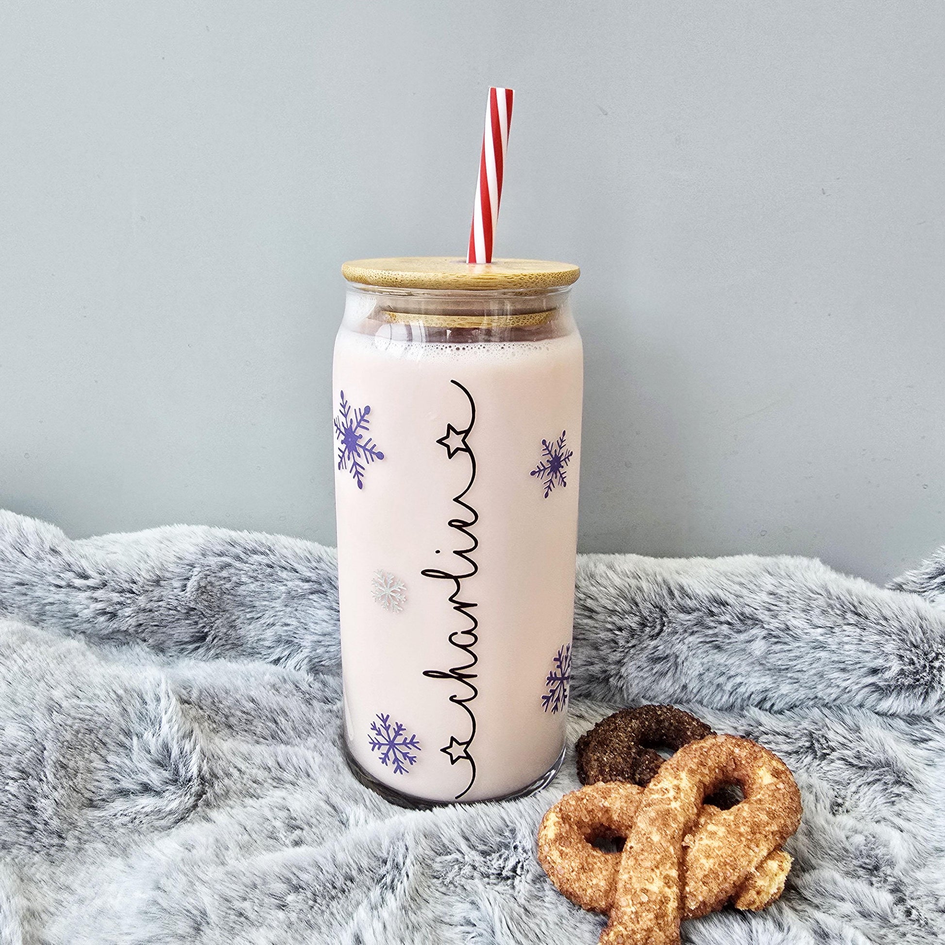 Personalised glass can drink holder with a bamboo suction lid and red and white reusable straw. Features silver snowflakes and blue snowflakes that colour change to purple when cold liquid is added to the glass. Personalised name along the side of the glass, available in glittery silver or black and features either a heart or stars at either end of the name.
