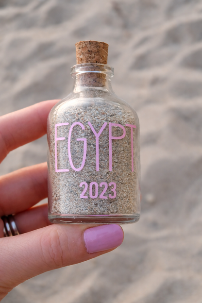 Small bottle with cork lid labeled 'Egypt 2023' held by a hand on a sandy background
