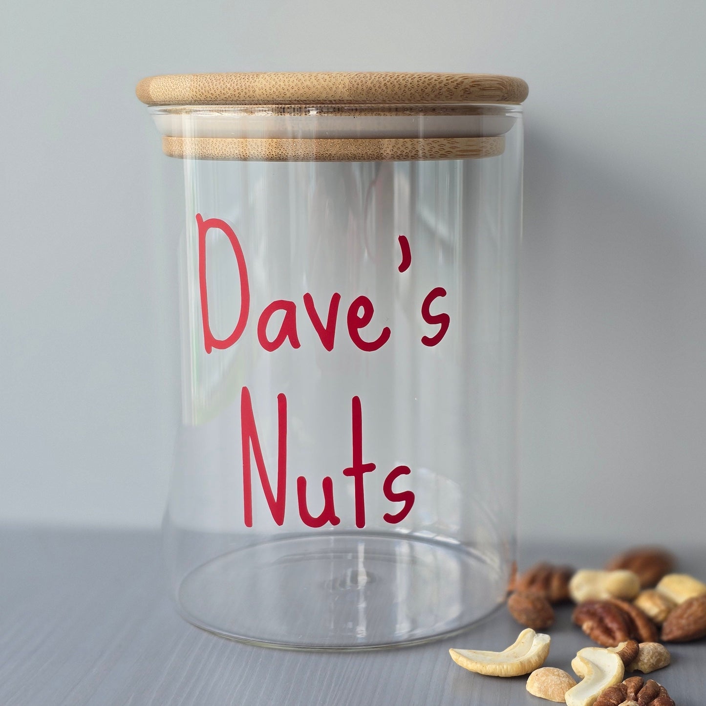 Personalised glass jar with wooden suction lids, labeled 'Dave's Nuts' on a grey surface with nuts around.