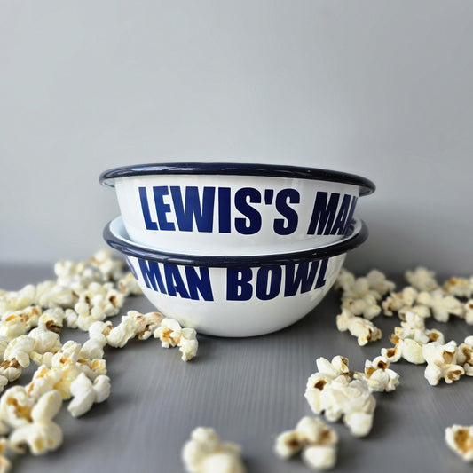 White enamel metal bowl with blue rim and text personalised with a name and 'MAN BOWL' surrounded by popcorn on a grey surface.