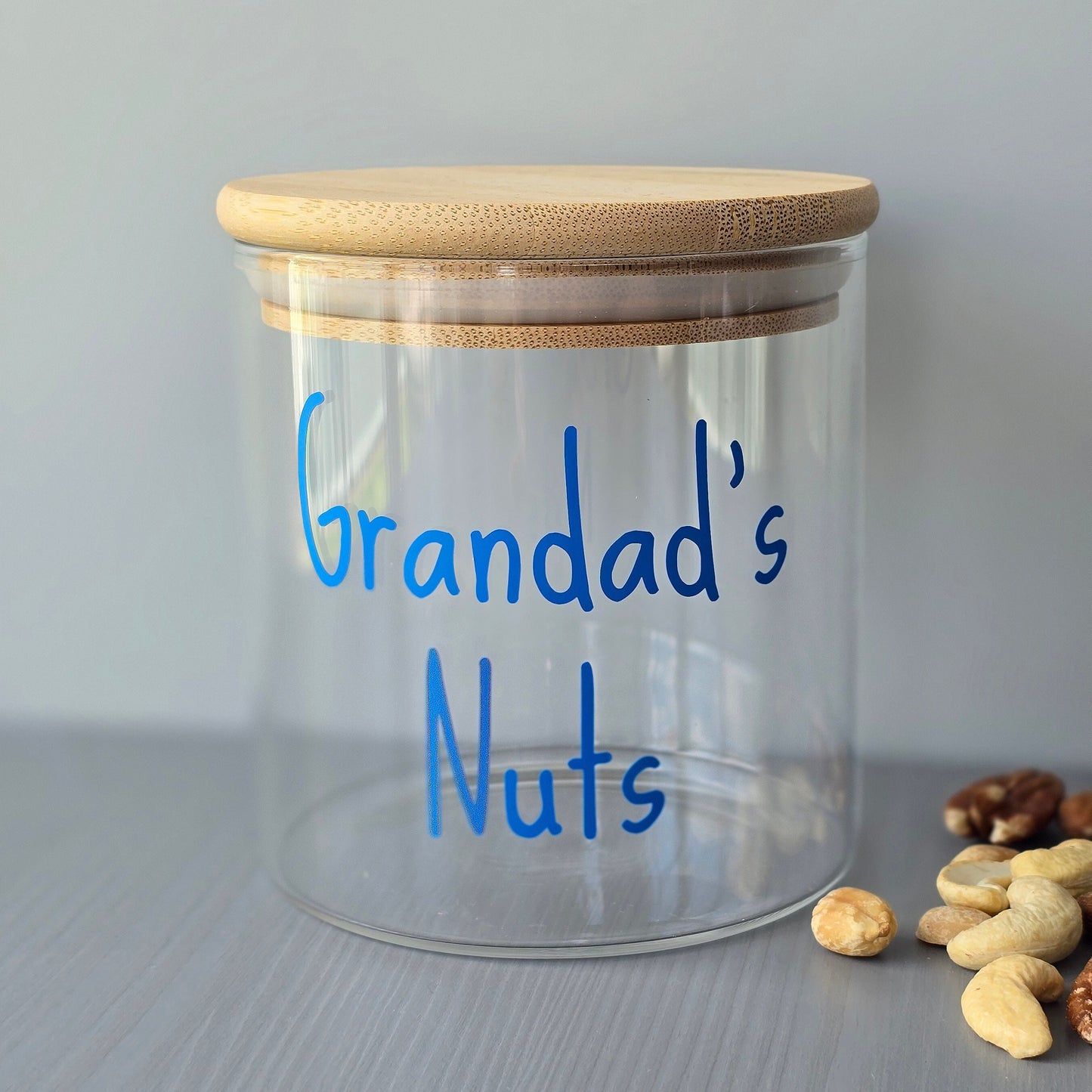 Personalised glass jar with wooden suction lids and 'Grandad's Nuts' on a grey surface.