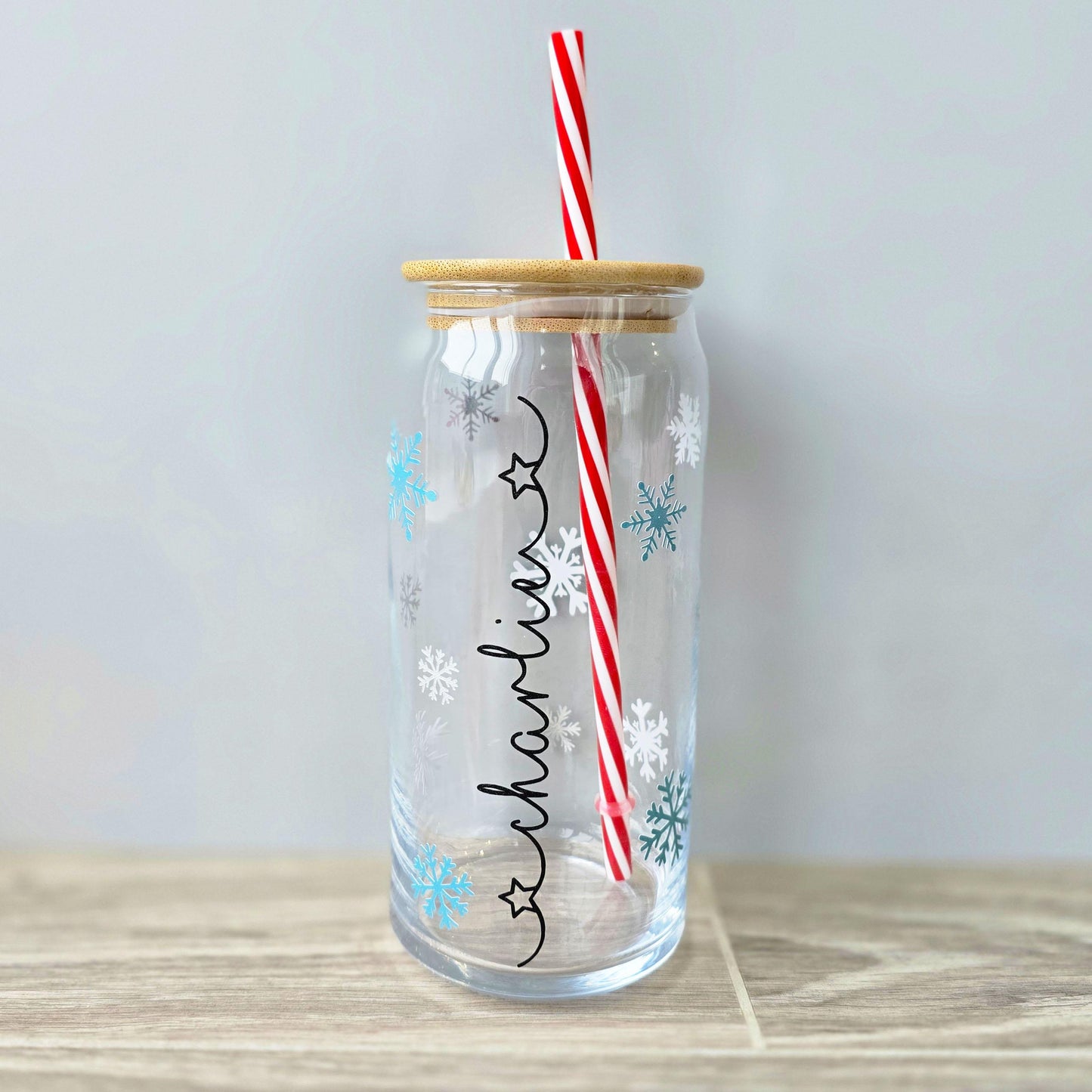 Personalised glass can drink holder with a bamboo suction lid and red and white reusable straw. Features silver snowflakes and blue snowflakes that colour change to purple when cold liquid is added to the glass. Personalised name along the side of the glass, available in glittery silver or black and features either a heart or stars at either end of the name.
