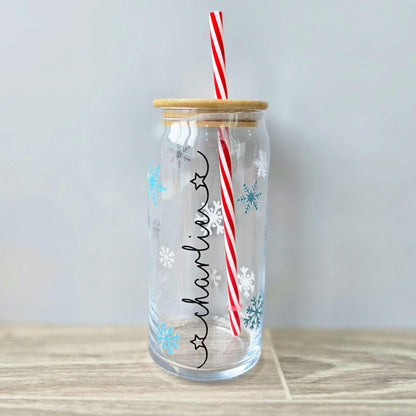 Personalised glass can drink holder with a bamboo suction lid and red and white reusable straw. Features silver snowflakes and blue snowflakes that colour change to purple when cold liquid is added to the glass. Personalised name along the side of the glass, available in glittery silver or black and features either a heart or stars at either end of the name.