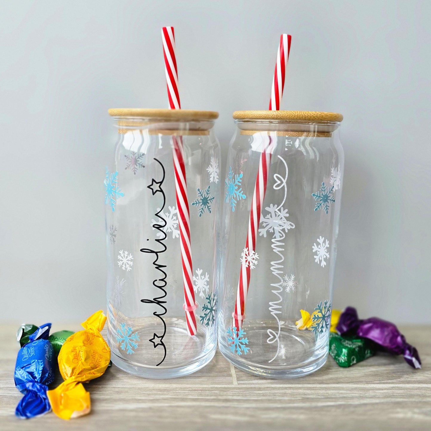 Personalised glass can drink holder with a bamboo suction lid and red and white reusable straw. Features silver snowflakes and blue snowflakes that colour change to purple when cold liquid is added to the glass. Personalised name along the side of the glass, available in glittery silver or black and features either a heart or stars at either end of the name.