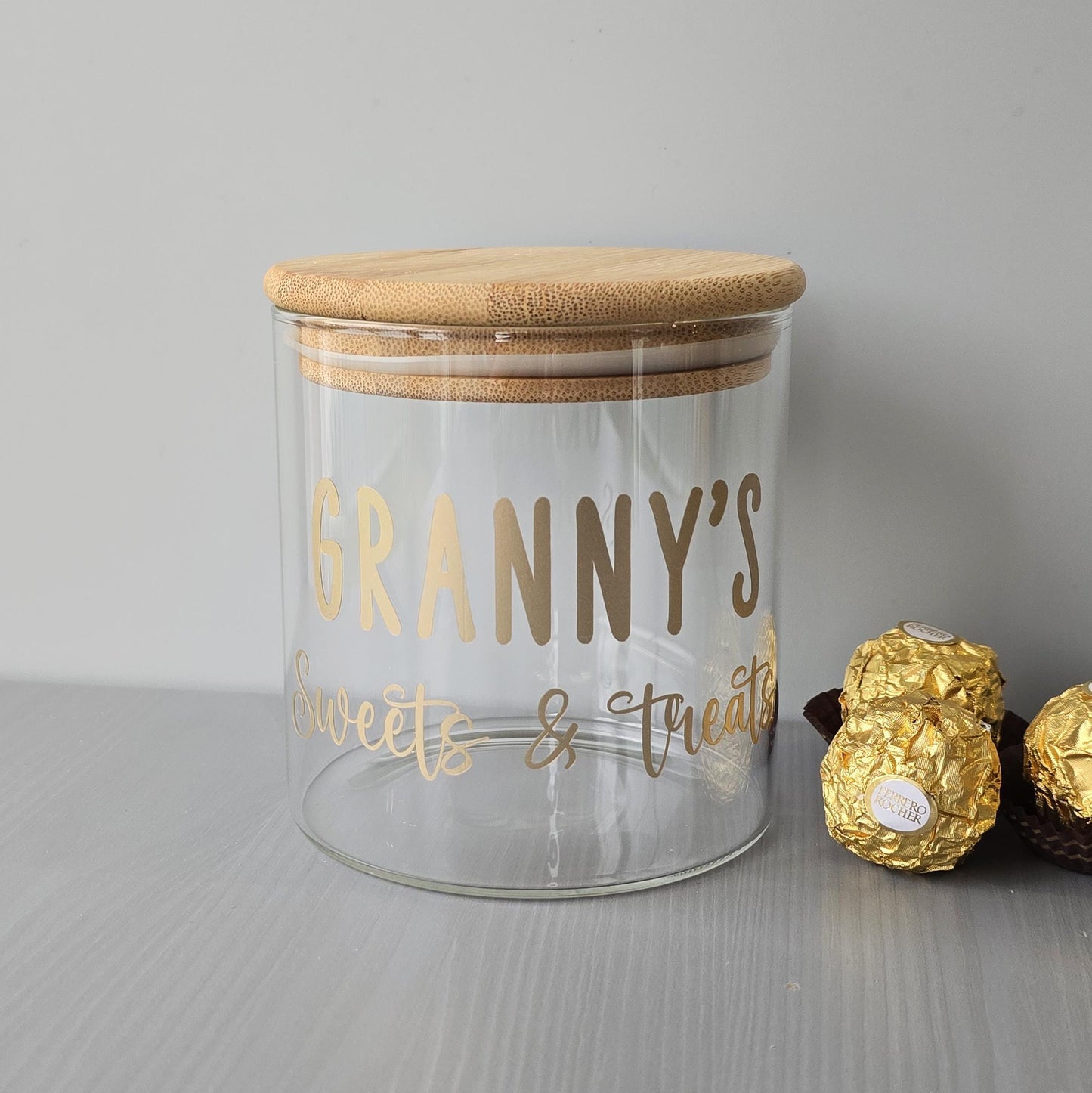 Glass jar with wooden lid labeled 'Granny's Sweets & Treats' on a gray surface with wrapped candies.