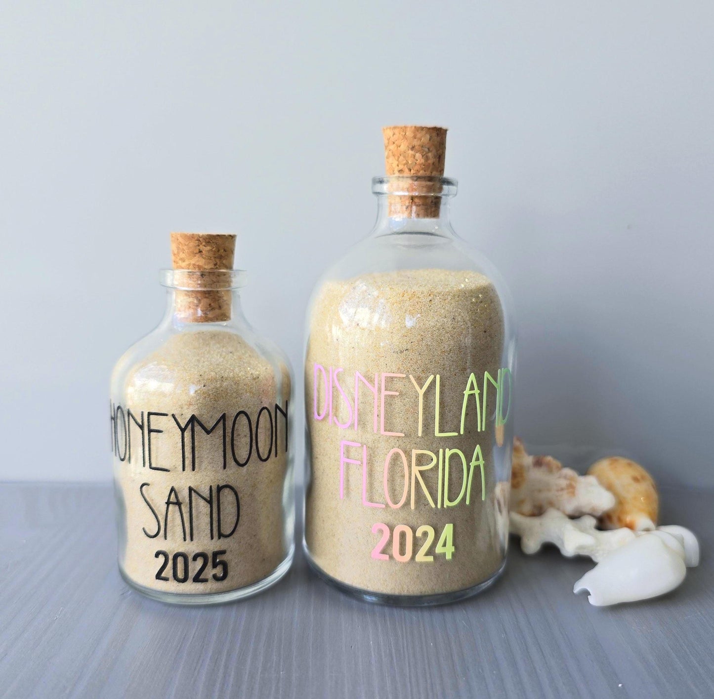 Two glass bottles filled with sand, labeled 'Honeymoon Sand 2025' and 'Disneyland Florida 2024', on a gray surface.