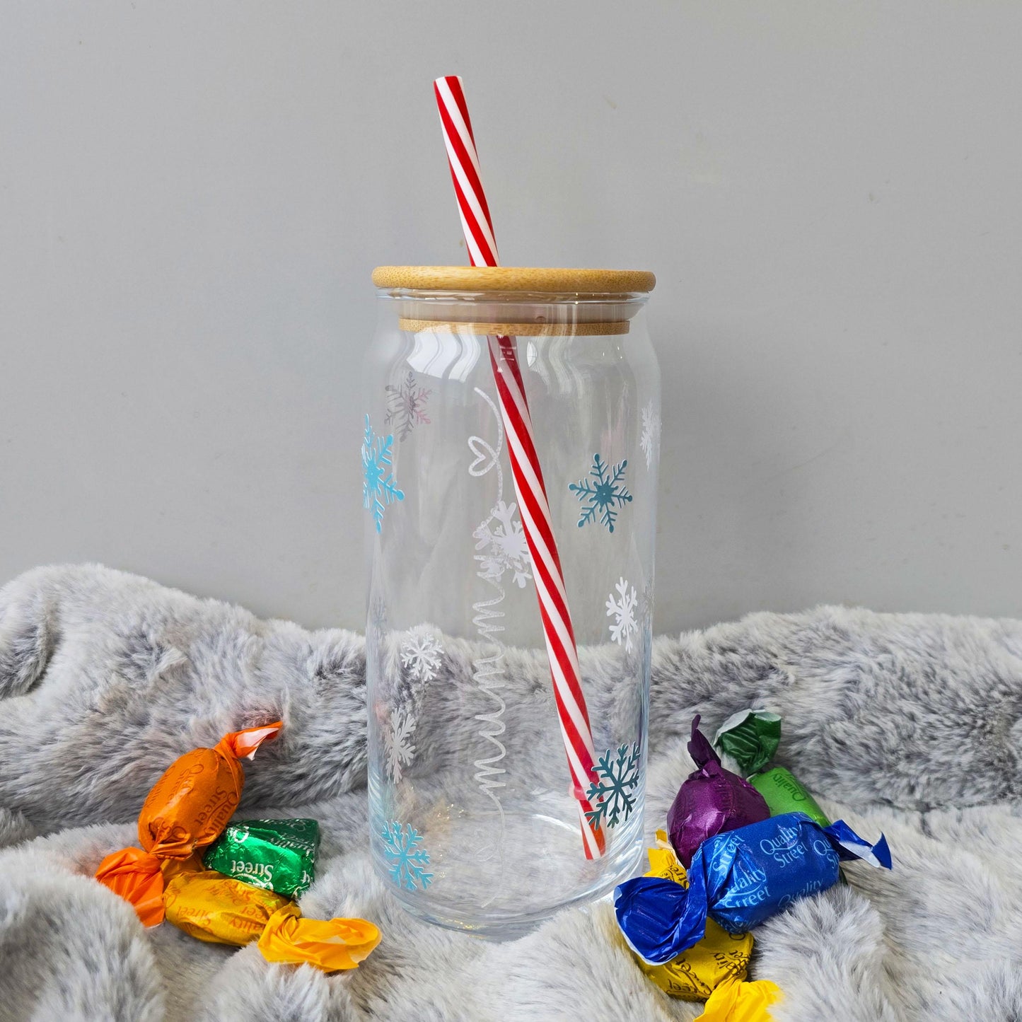 Personalised glass can drink holder with a bamboo suction lid and red and white reusable straw. Features silver snowflakes and blue snowflakes that colour change to purple when cold liquid is added to the glass. Personalised name along the side of the glass, available in glittery silver or black and features either a heart or stars at either end of the name.