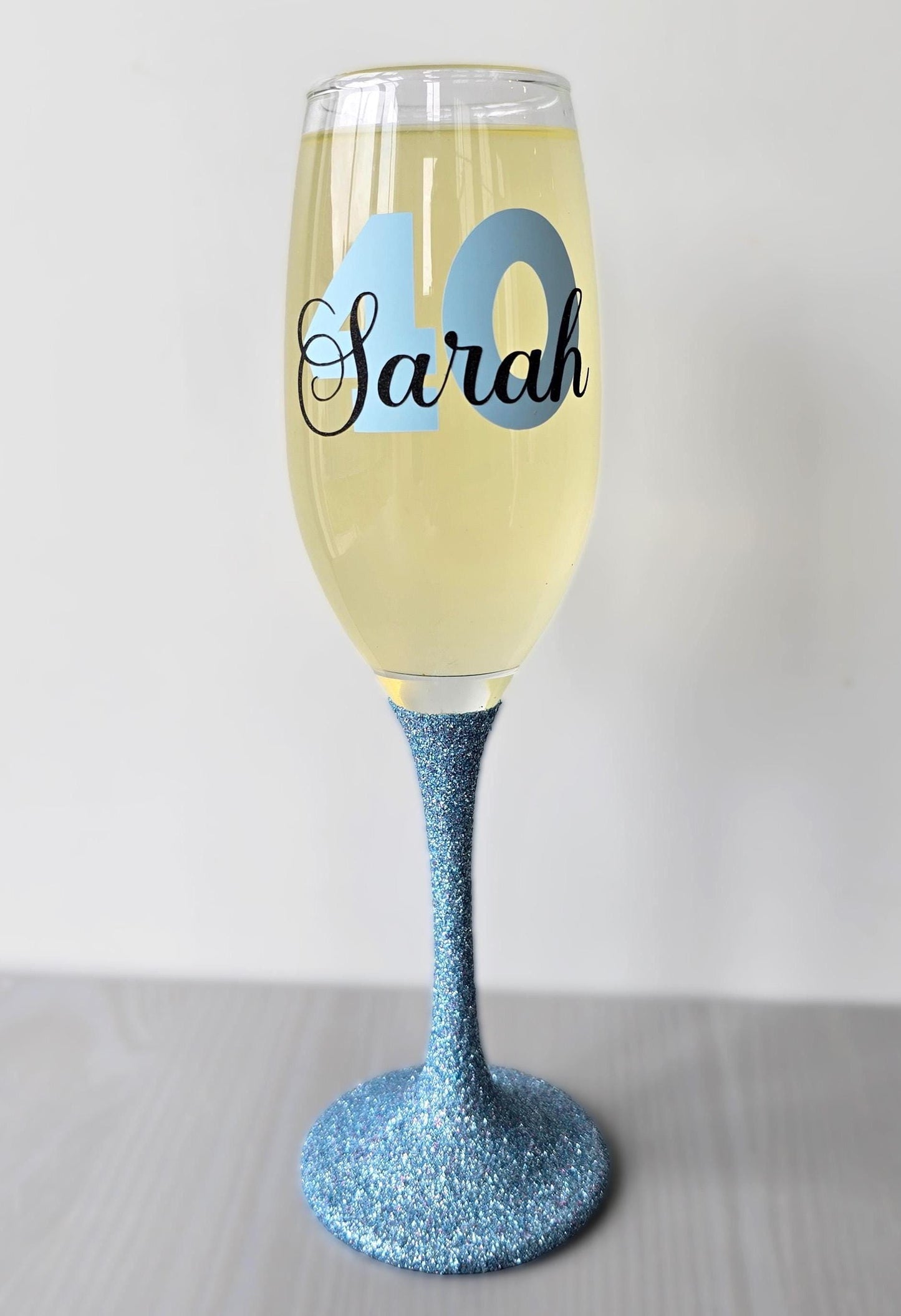 Personalised champagne flute or prosecco glass with glitter stem and with a birthday age number in block, glitter or metallic vinyl and a name in black decorative font through the number