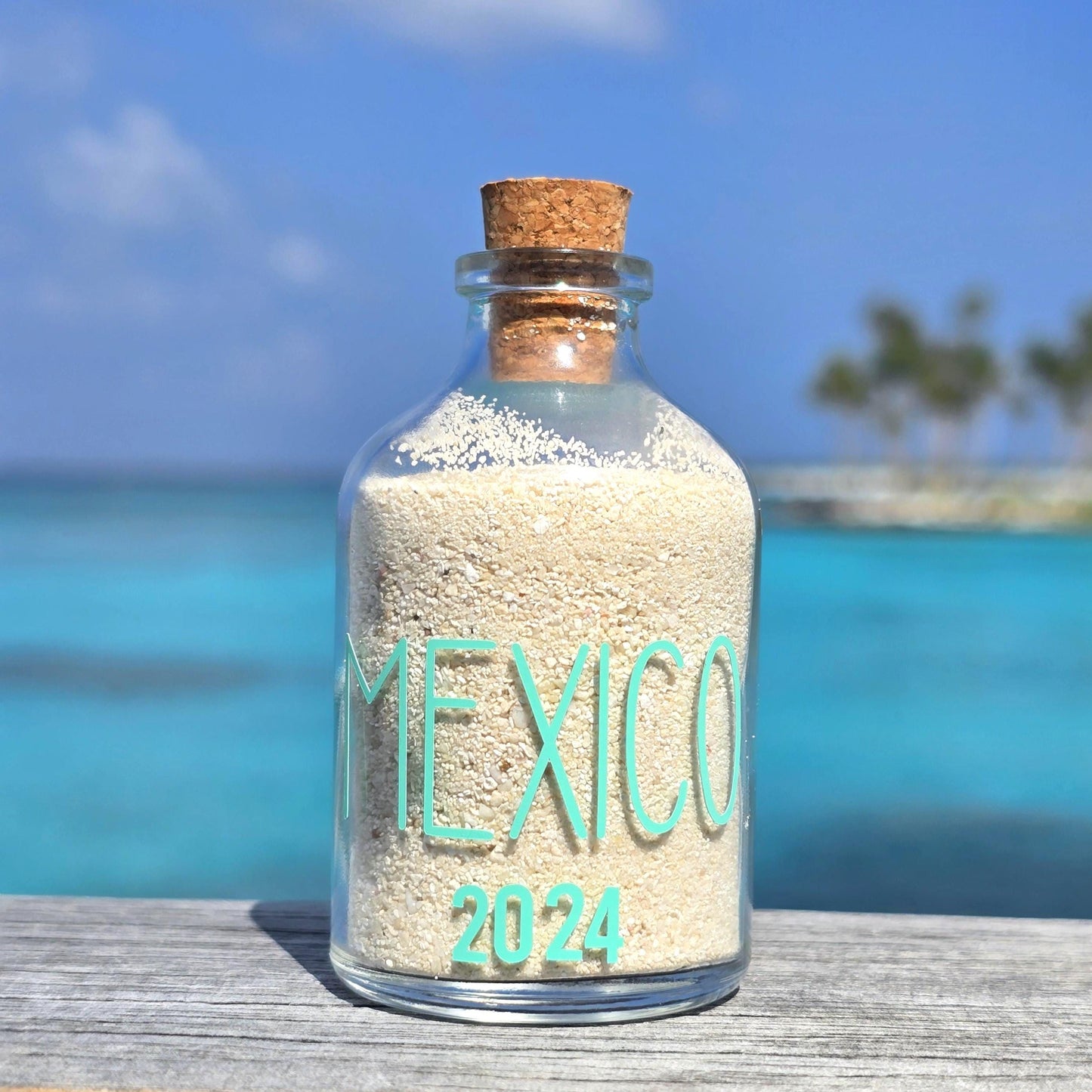 Glass bottle with cork lid containing sand labeled 'Mexico 2024' against a beach background